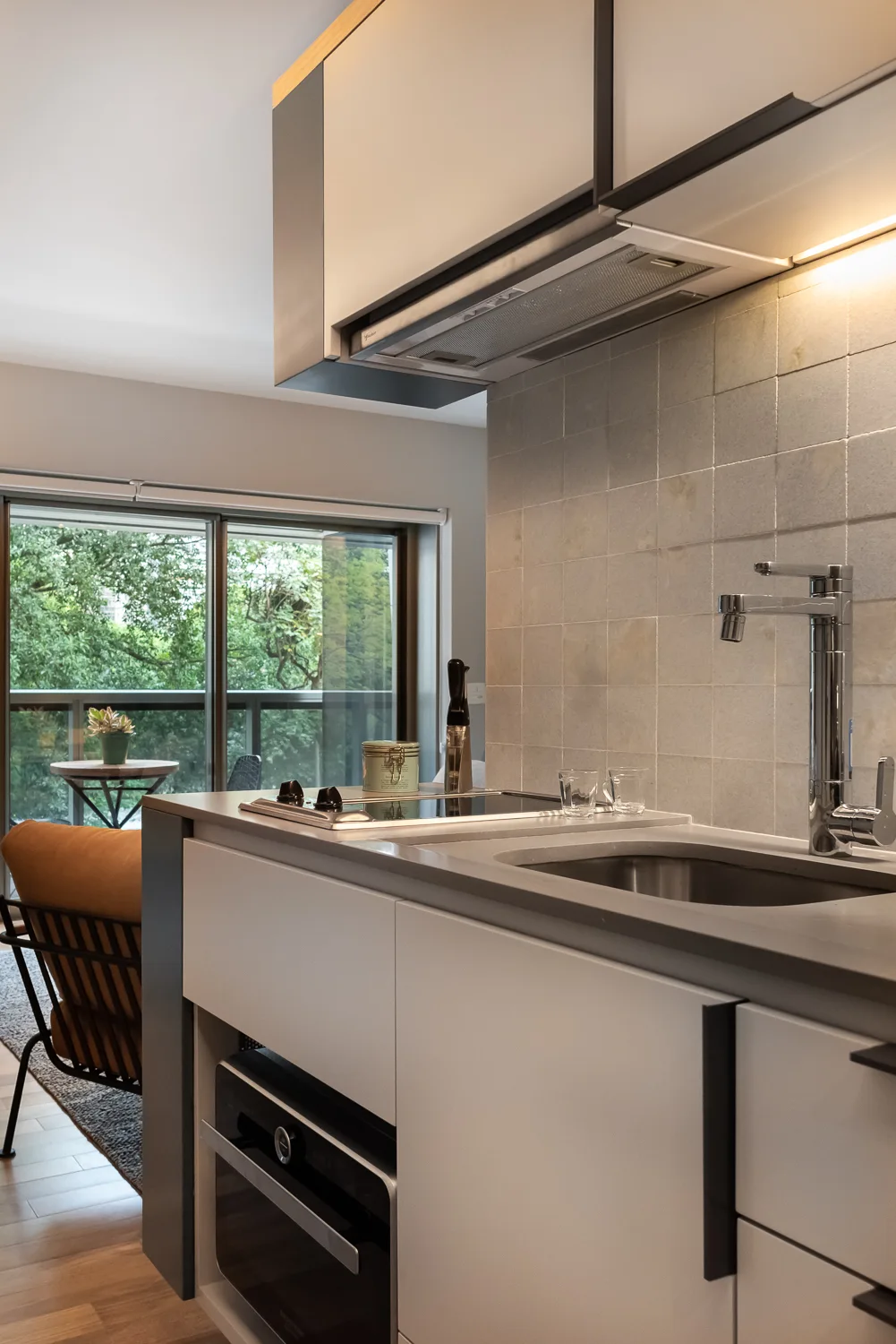 A modern compact kitchen featuring white flat-panel cabinets, a grey square-tile backsplash, an integrated sink, and an under-cabinet light.