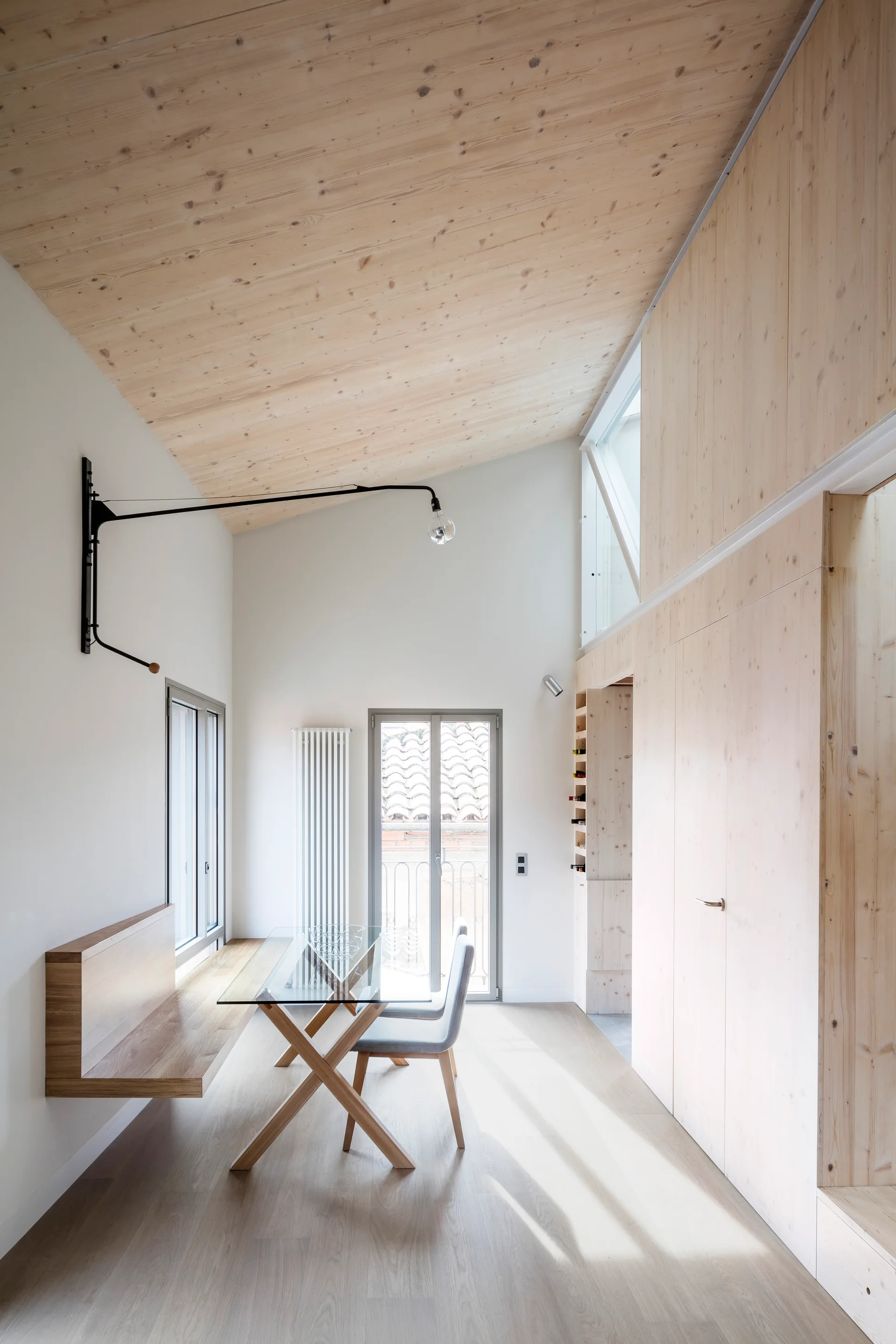 A minimalist dining room features a glass-top table, a floating wooden bench, a sloped light-wood ceiling, and matching integrated wall cabinetry.