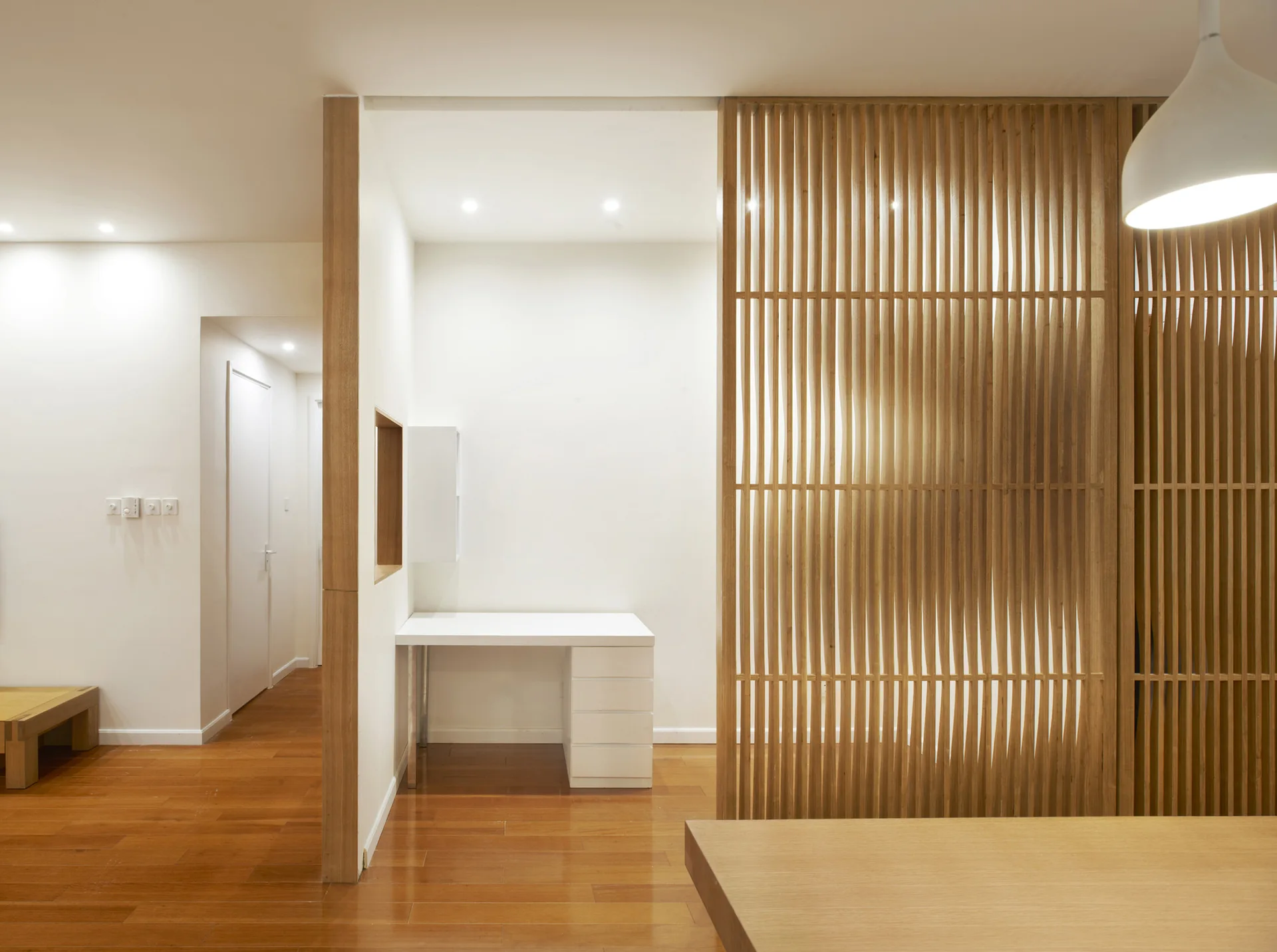 A minimalist home office features a white writing desk in a recessed nook separated from the main room by a vertical wooden slatted screen.