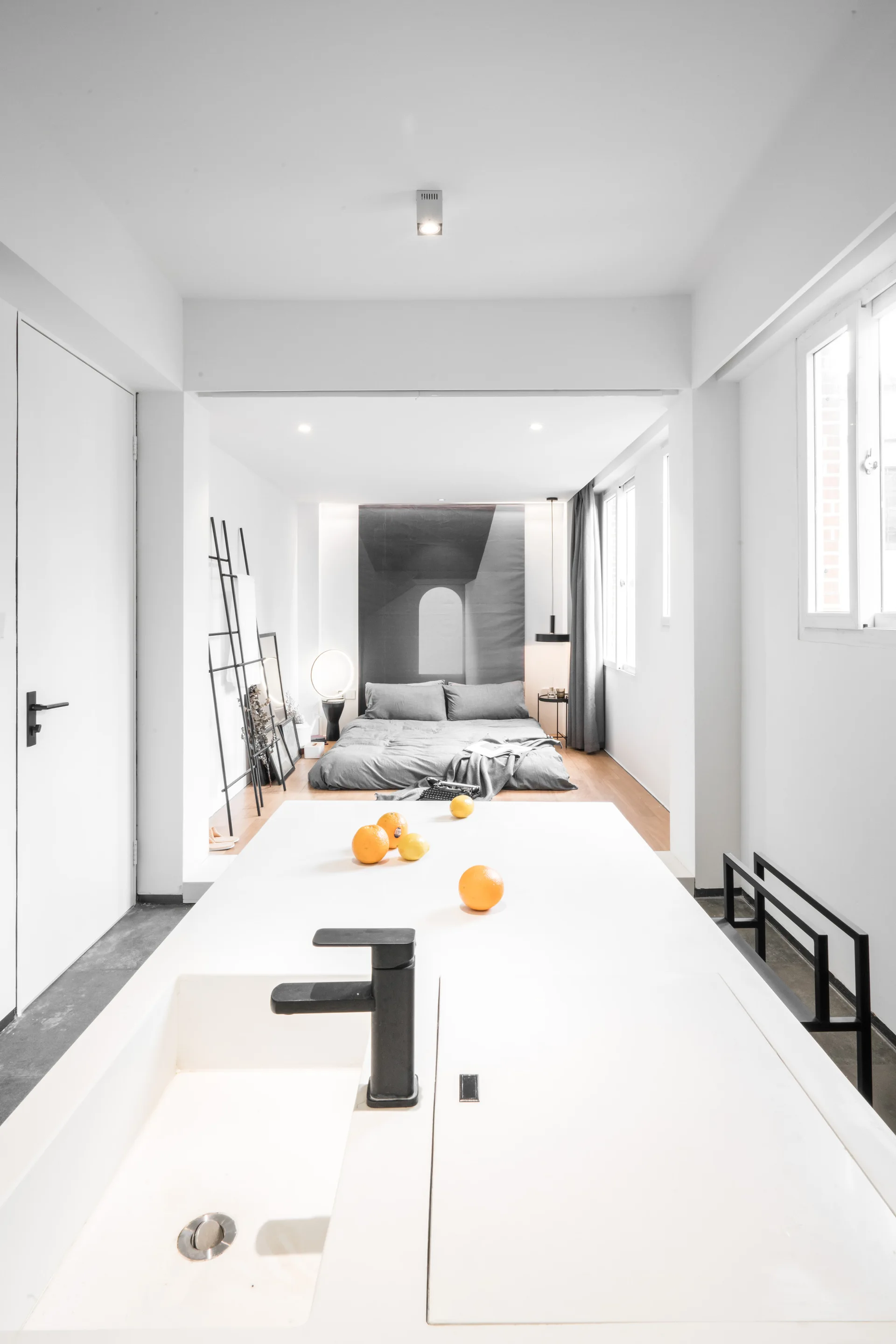 A minimalist studio apartment features a white kitchen island in the foreground and a sunken bedroom with a low grey bed in the background.