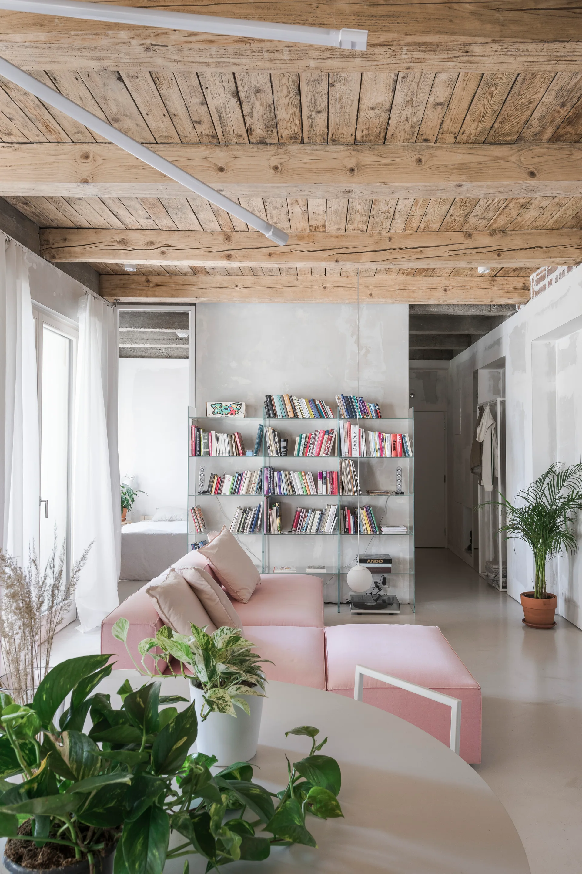 A modern living room features a pink sectional sofa, a tall glass bookshelf, and a rustic exposed wood beam ceiling.