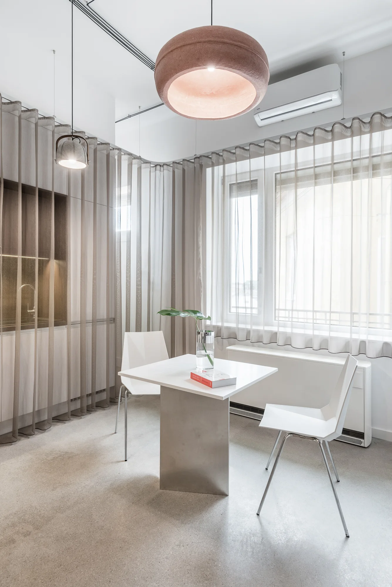 A minimalist dining nook features a white square table and two chairs set against a window and enclosed by a curved sheer curtain.