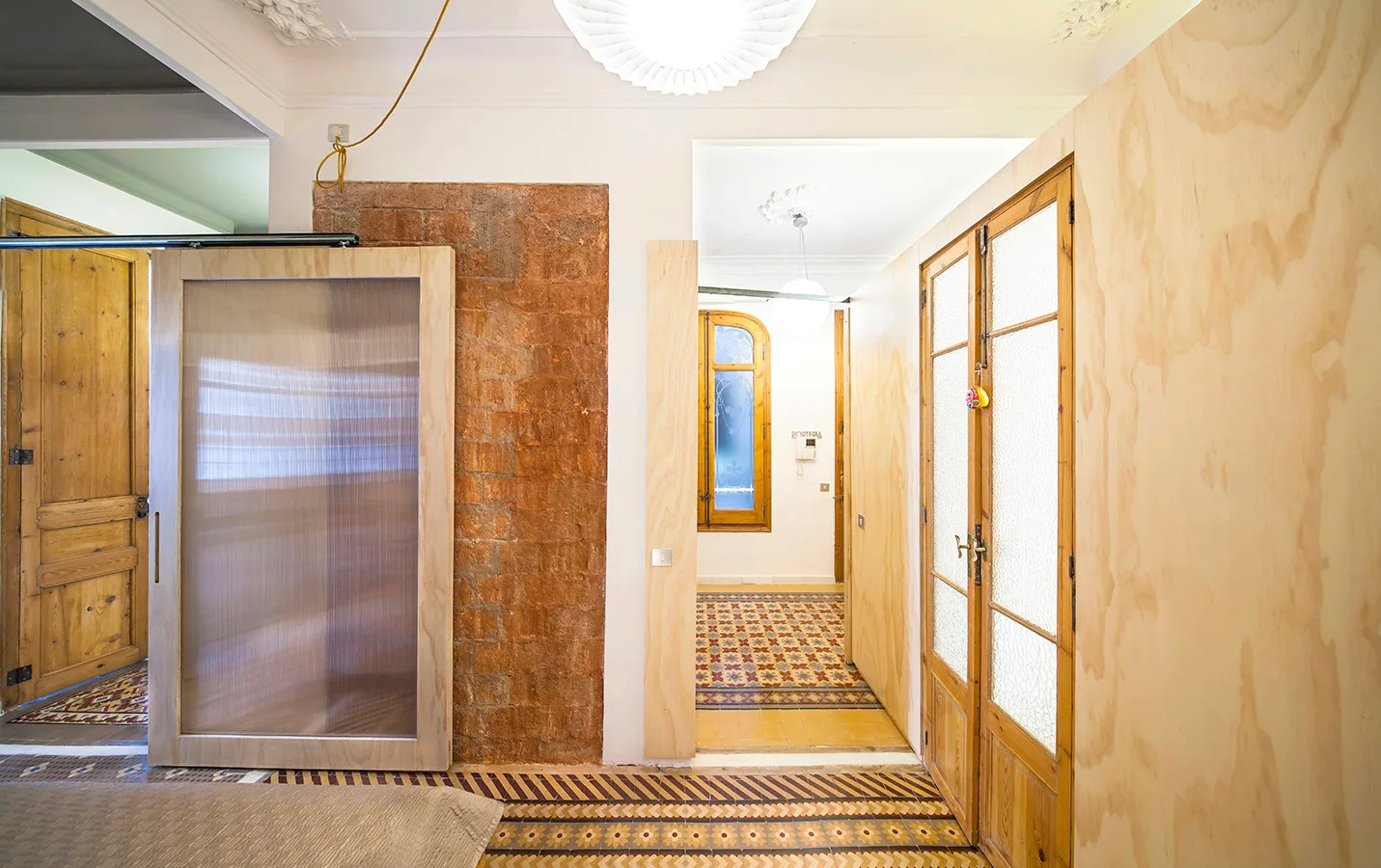 An interior hallway features light plywood wall paneling, a sliding door with a translucent corrugated panel, and a patterned mosaic tile floor.