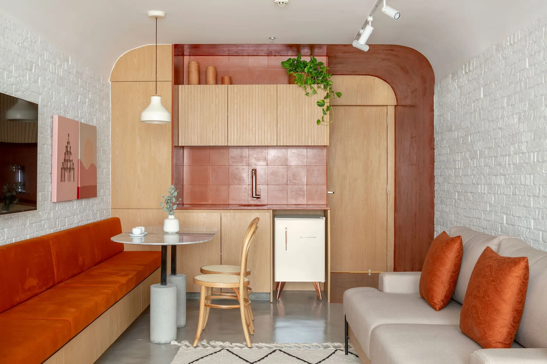 A compact studio apartment kitchenette featuring a painted terra cotta accent arch, light wood cabinets, and an orange banquette.