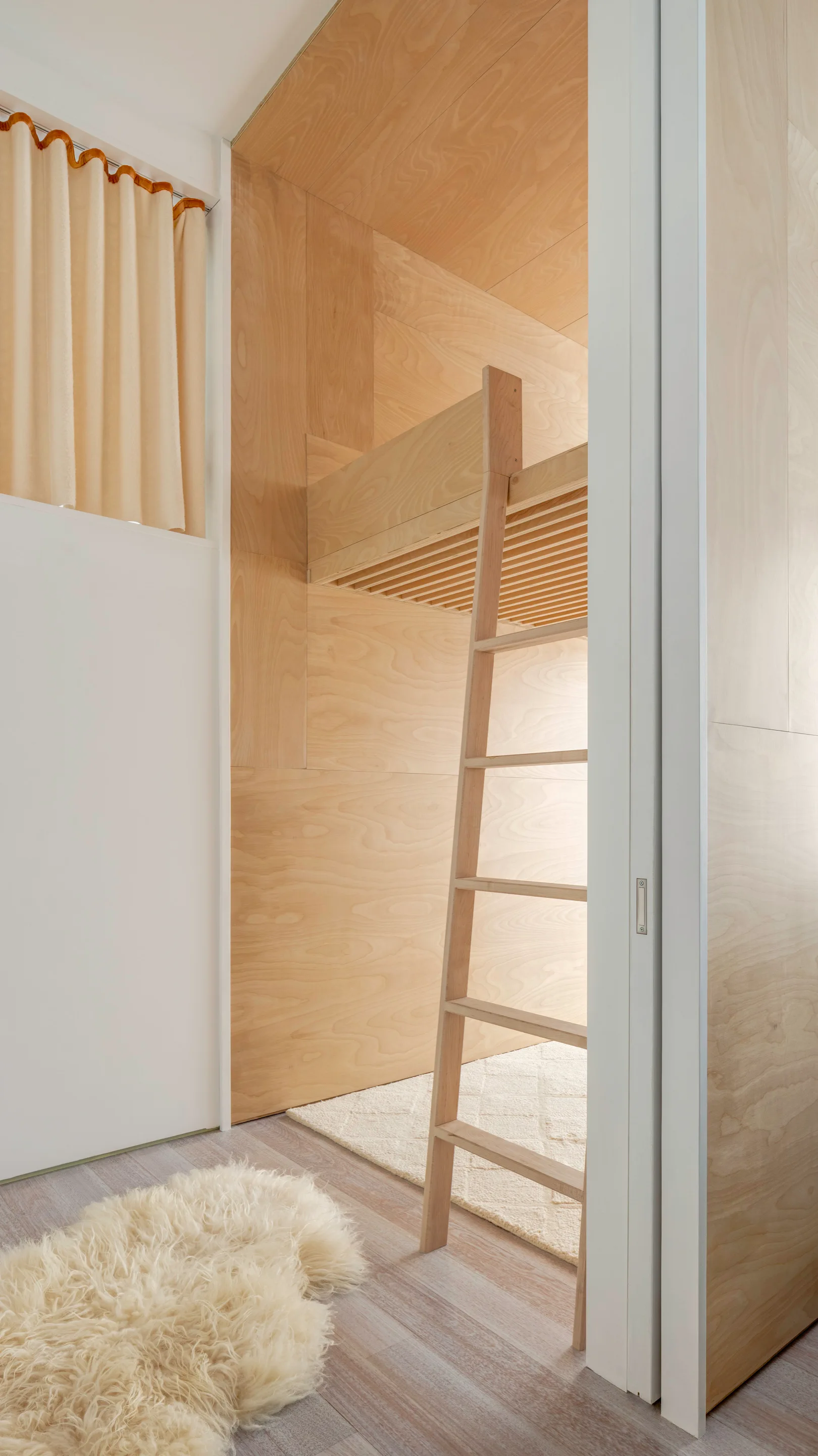 A built-in plywood loft bed with a wooden ladder sits inside a room with matching plywood wall paneling and a white sliding door.