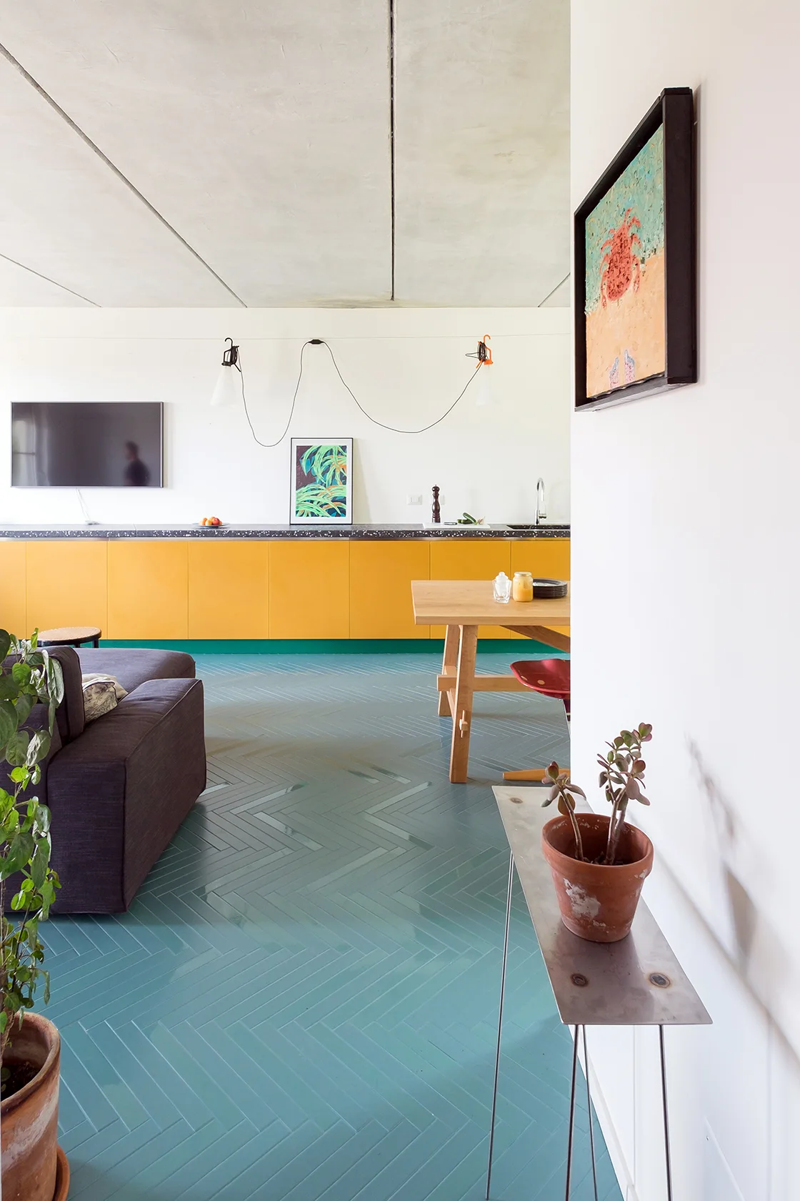 An open-plan room features a teal herringbone tile floor, bright yellow kitchen cabinets, a wooden dining table, and a grey sofa under a concrete ceiling.