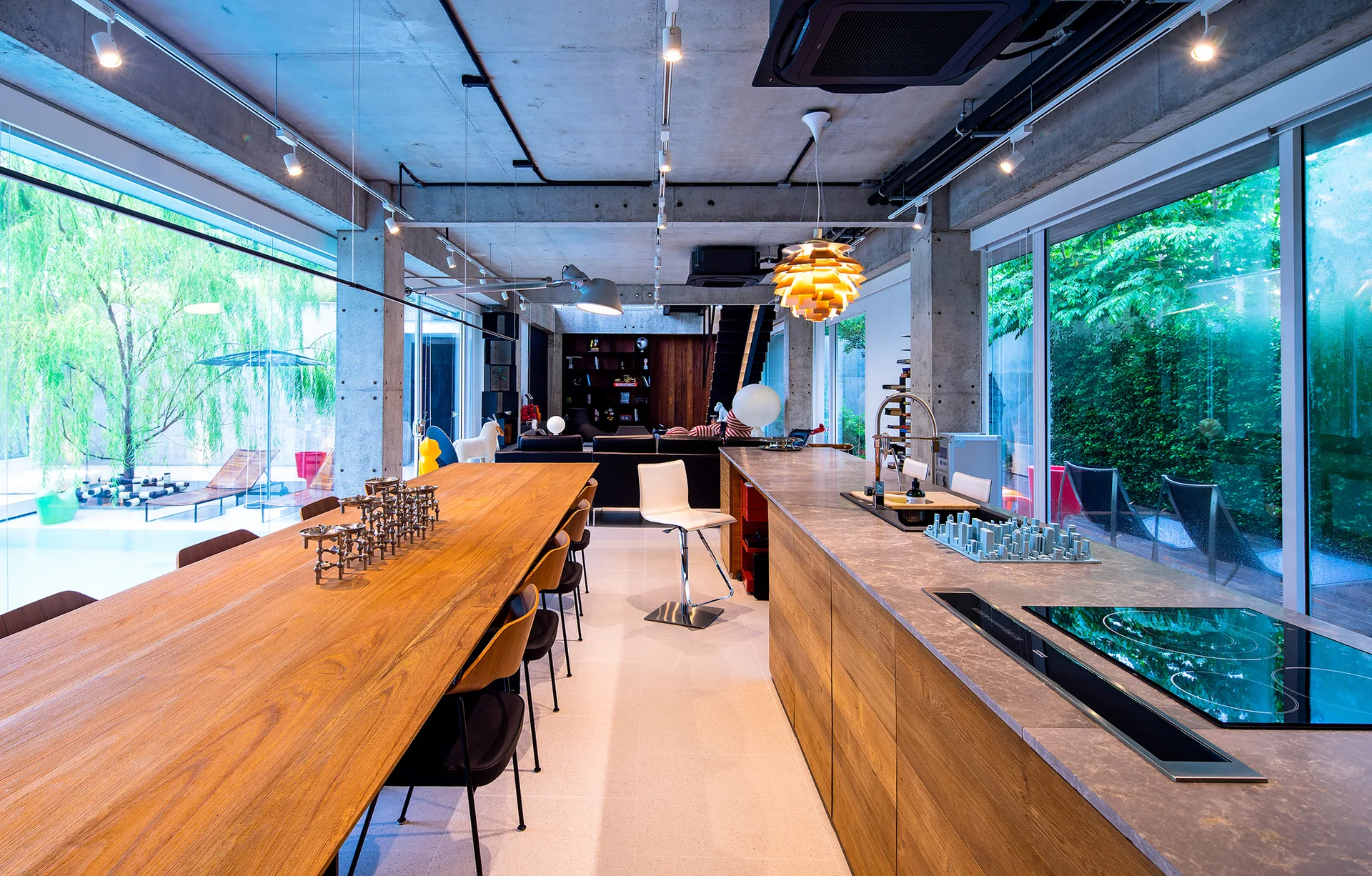 An industrial kitchen with raw concrete columns, a long wood dining table, a stone kitchen island, and glass walls looking out to a green garden.