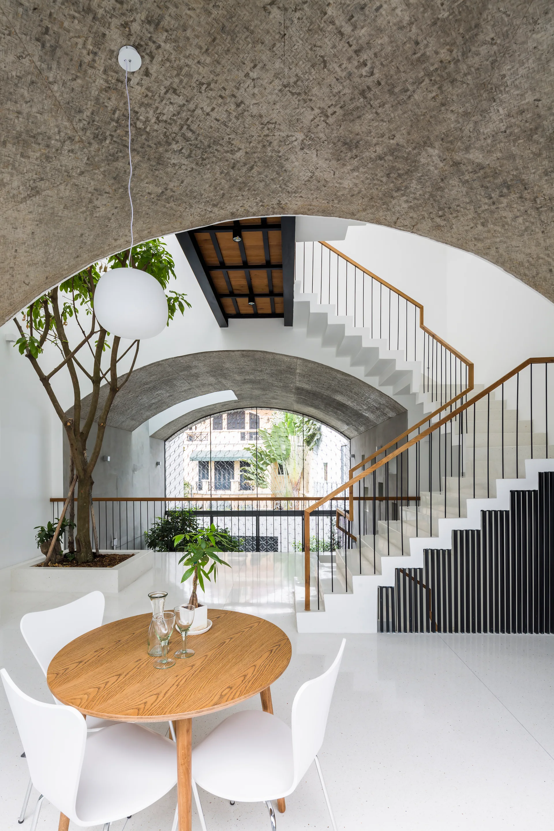 A modern dining area with a round wood table, white chairs, a sculptural white staircase, and an indoor tree beneath a textured concrete vault.