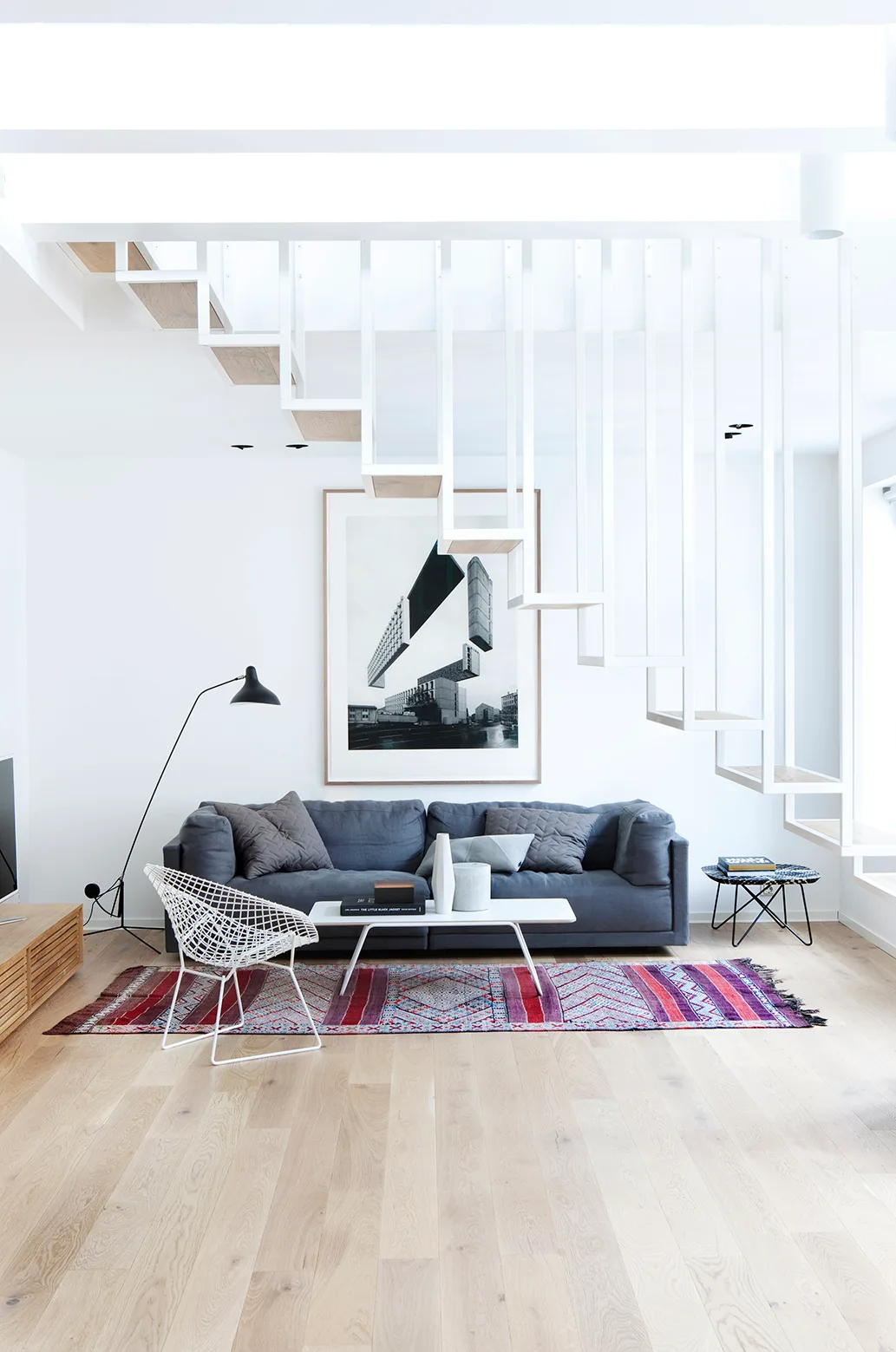 A modern living room features a grey sofa, a colorful patterned rug, and a white hanging staircase above light wood floors.