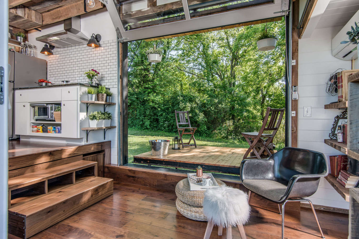 A compact interior with wood flooring, a raised kitchen, and a glass garage door rolled open to a wooden deck surrounded by trees.