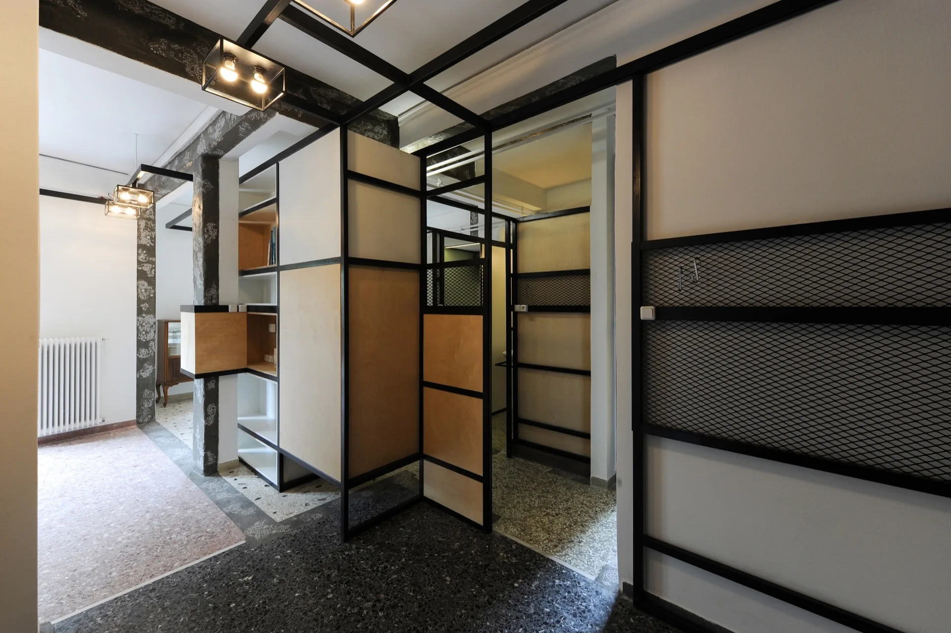 A modernist hallway features black metal and plywood partitions, integrated shelving, and dark terrazzo flooring under minimalist ceiling lights.