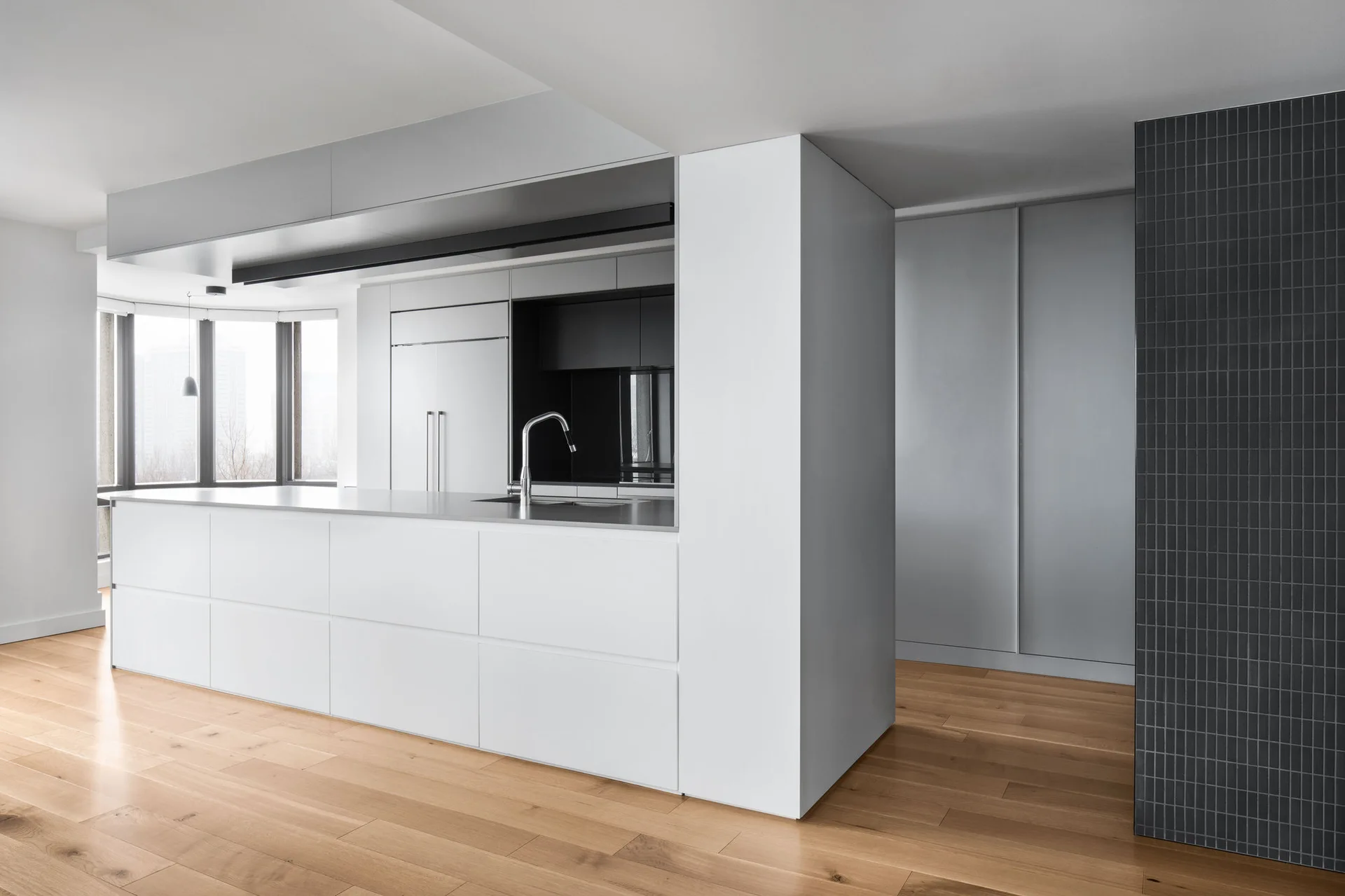 A minimalist kitchen features a large white island, integrated appliances, light hardwood floors, and a dark grey tiled accent column.