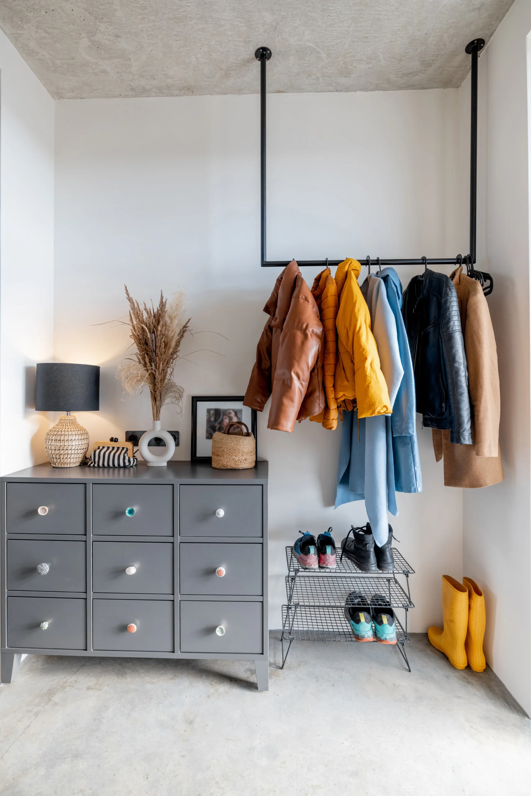 A modern entryway features a grey chest of drawers with decorative objects next to a ceiling-mounted black metal coat rack and shoe organizer.