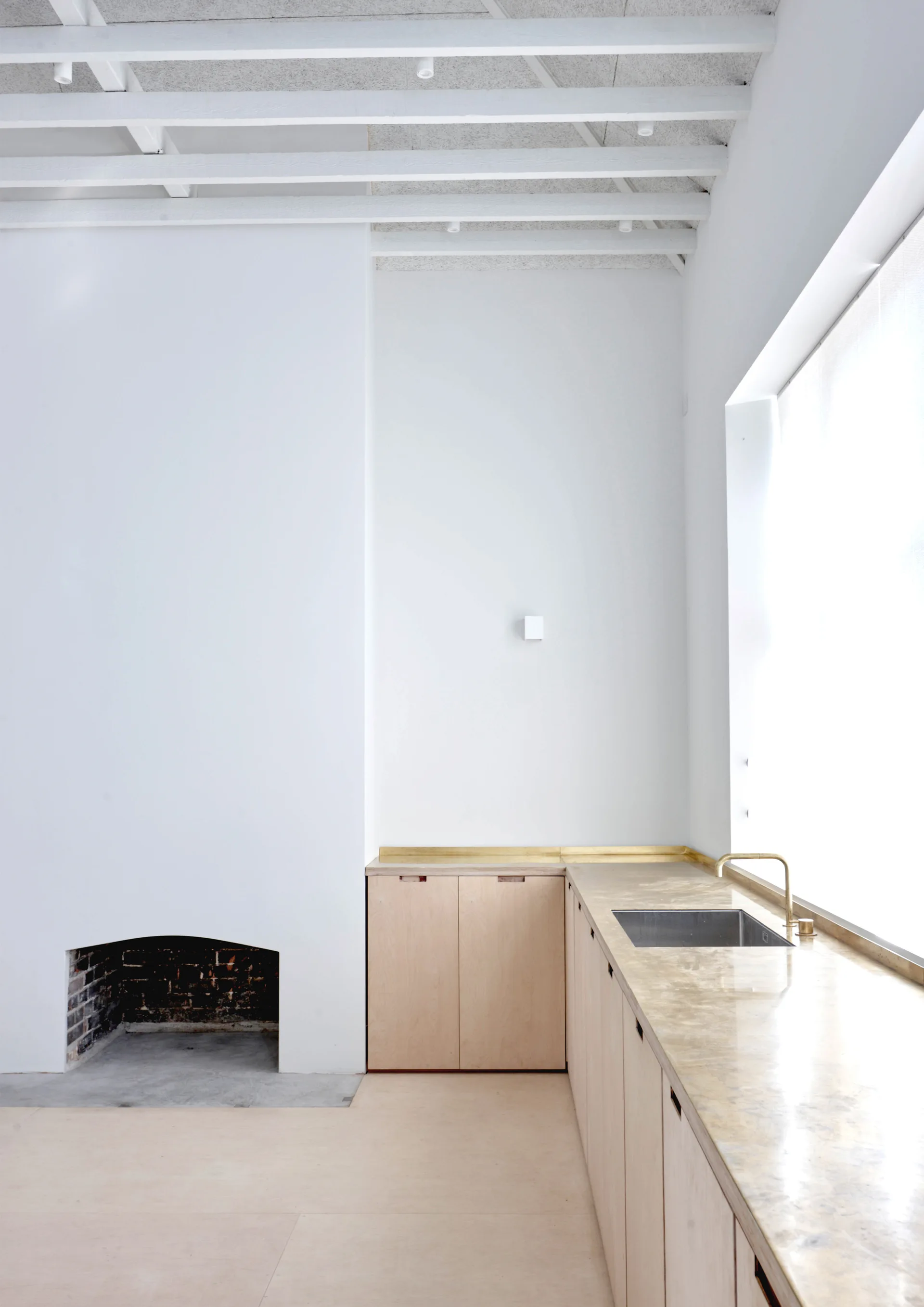 A minimalist kitchen features light wood plywood cabinets, a long brass countertop with a sink, white painted walls, and an arched fireplace.