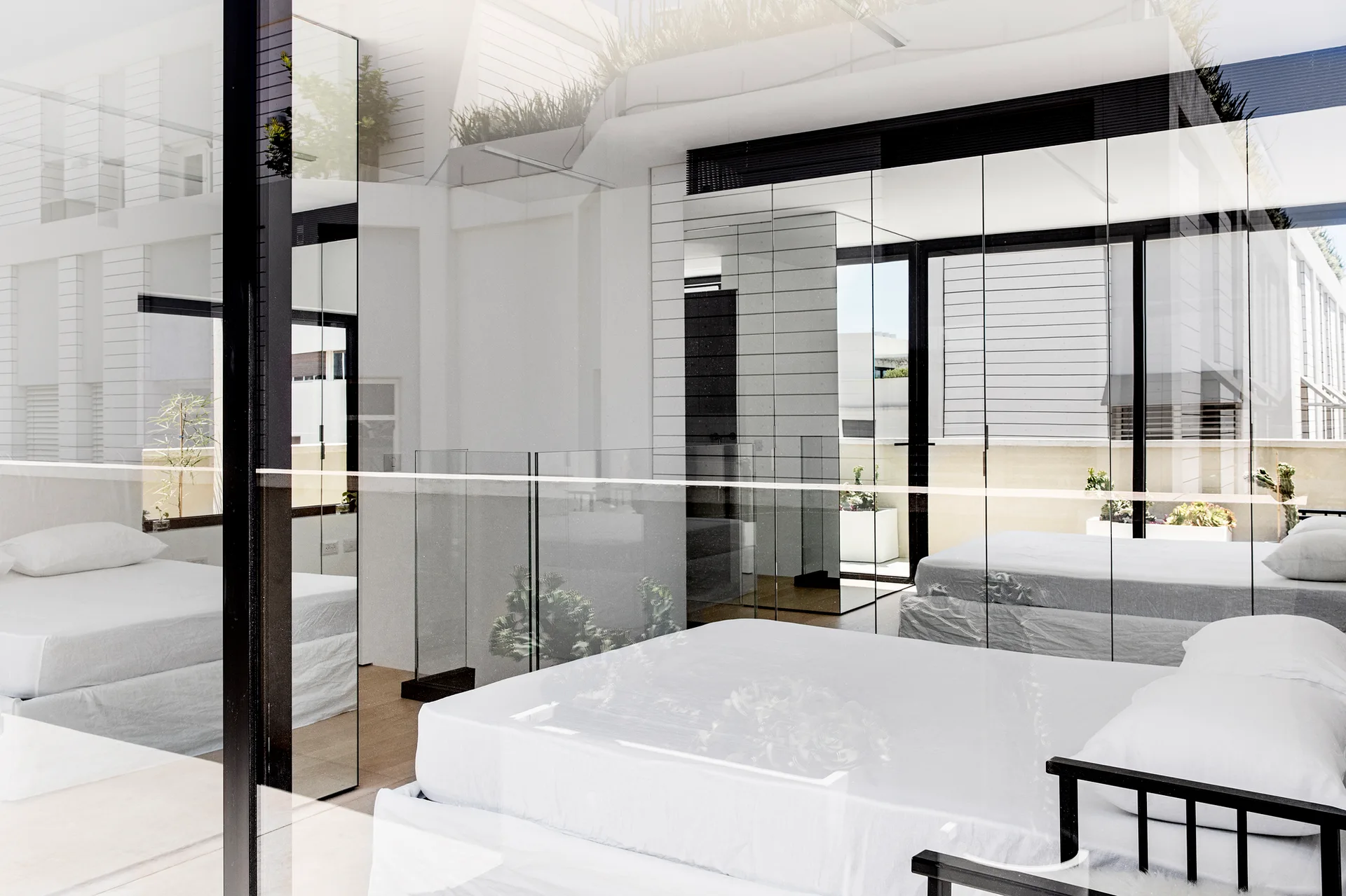 A minimalist bedroom features a white platform bed reflected in large mirrored wall panels next to a transparent glass partition.