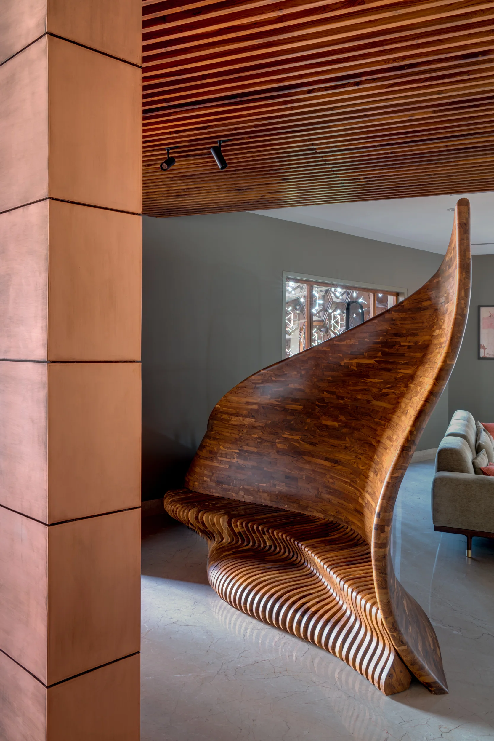 A sculptural curved wooden bench sits next to a square copper column under a slatted timber ceiling in a contemporary room.