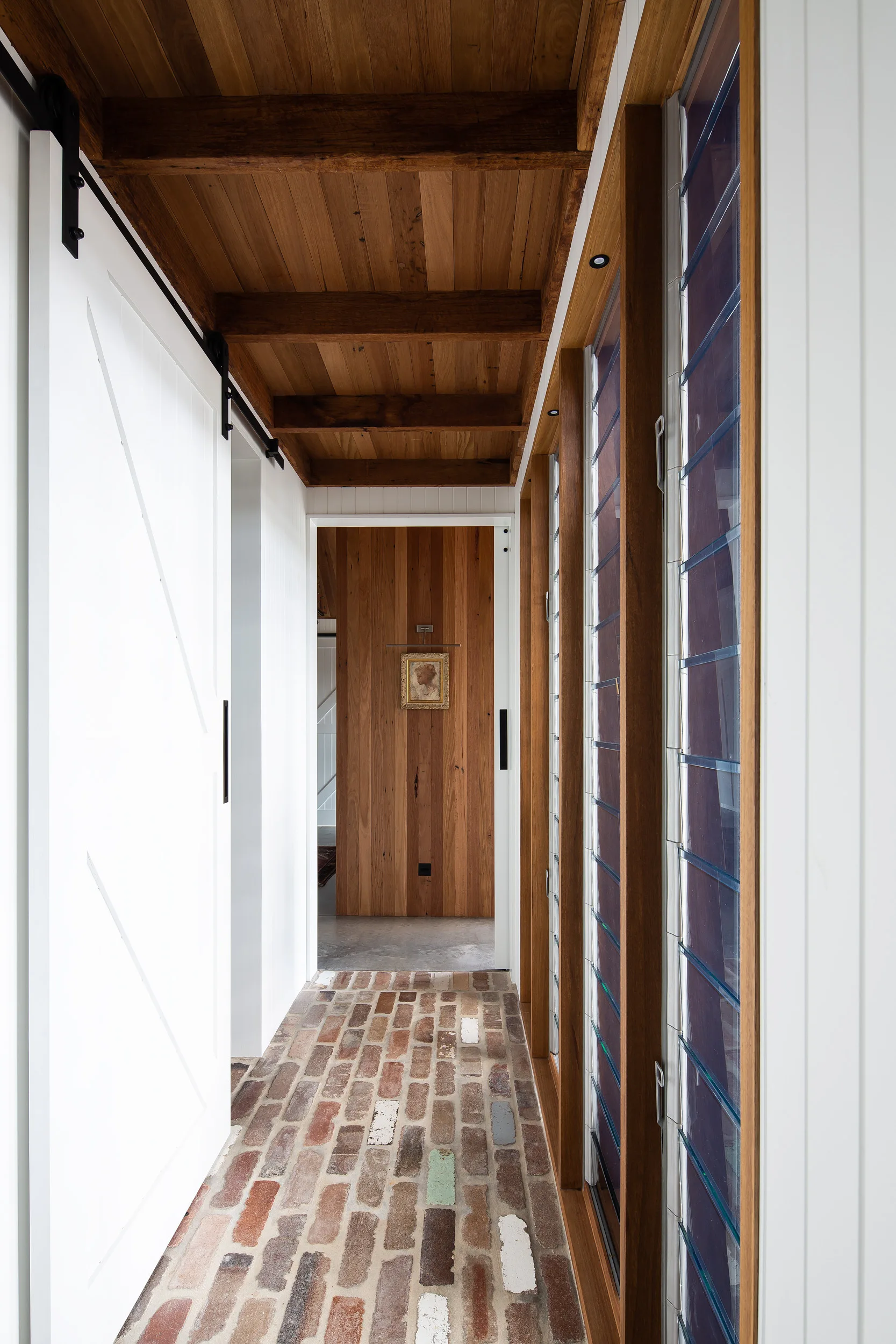 A narrow hallway features a multi-toned weathered brick floor, a white sliding barn door, louvered glass windows, and a warm wood-planked ceiling.
