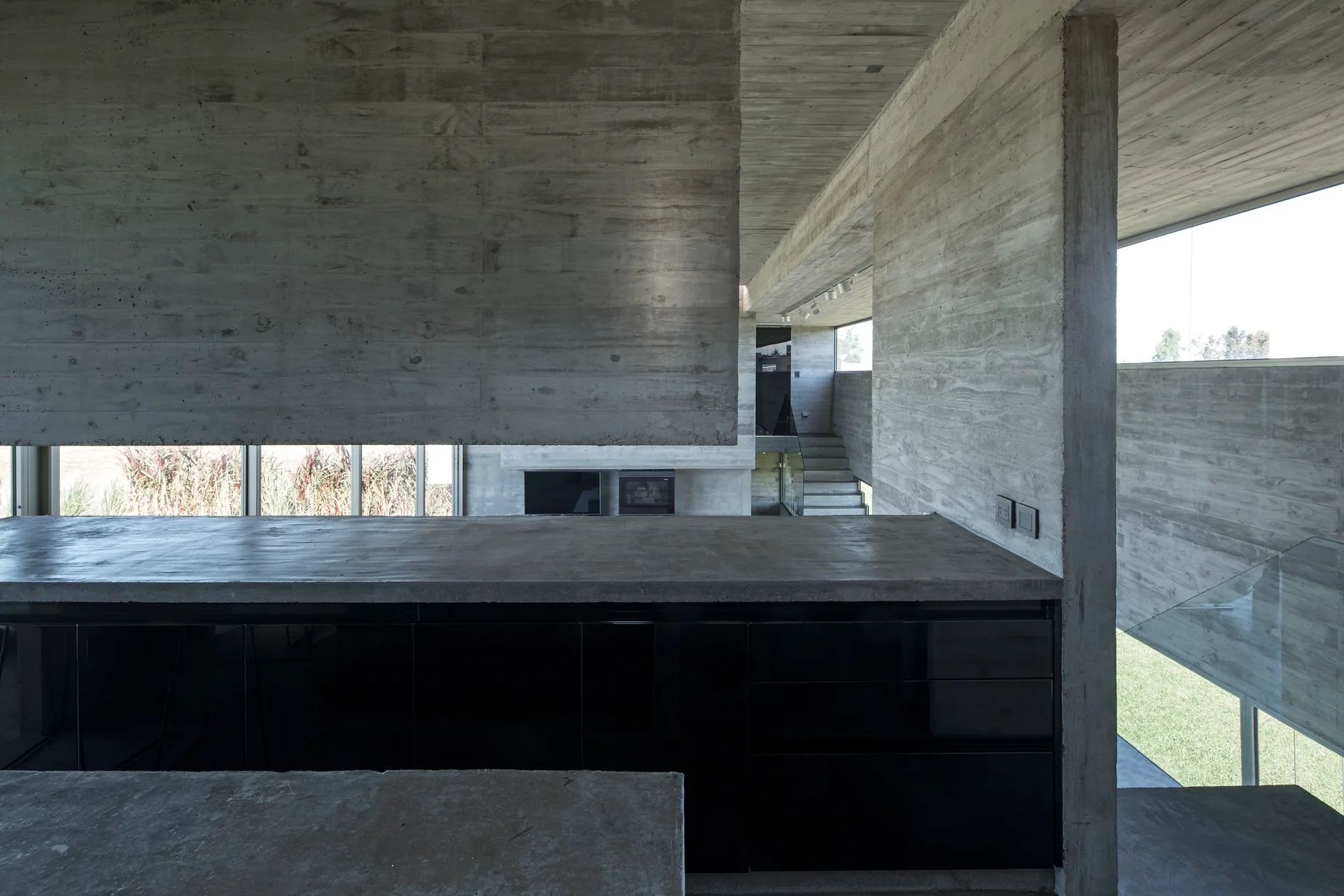 A minimalist kitchen features raw board-formed concrete walls and ceilings paired with a concrete island and high-gloss black cabinets.