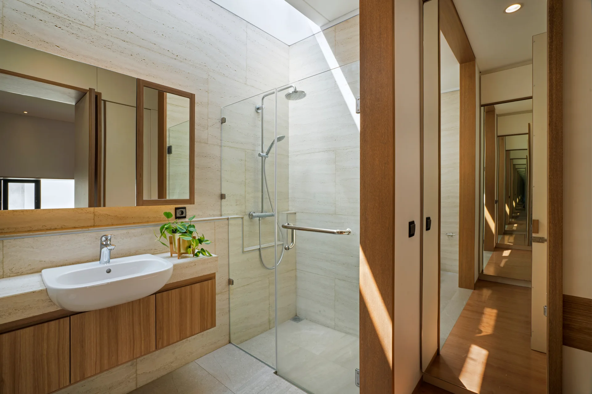 A modern beige bathroom features a wood-toned vanity with a white sink next to a glass walk-in shower illuminated by a large overhead skylight.