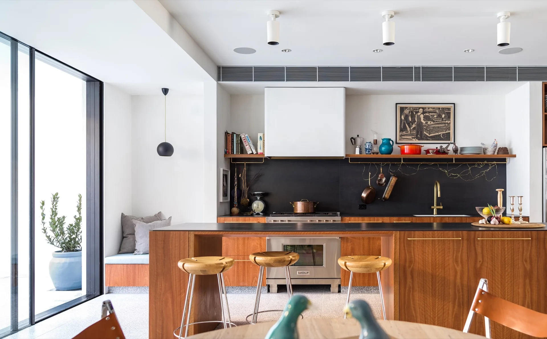 A modern kitchen featuring warm wood cabinetry, a matte black backsplash, three wooden bar stools, and a large central kitchen island.