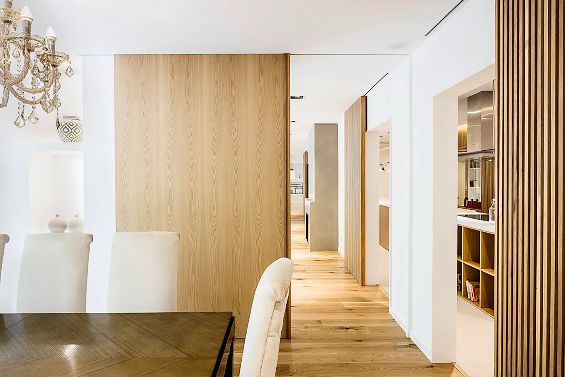 A modern dining room with cream upholstered chairs and a dark wood table features a large light oak wood partition wall opening to a bright hallway.