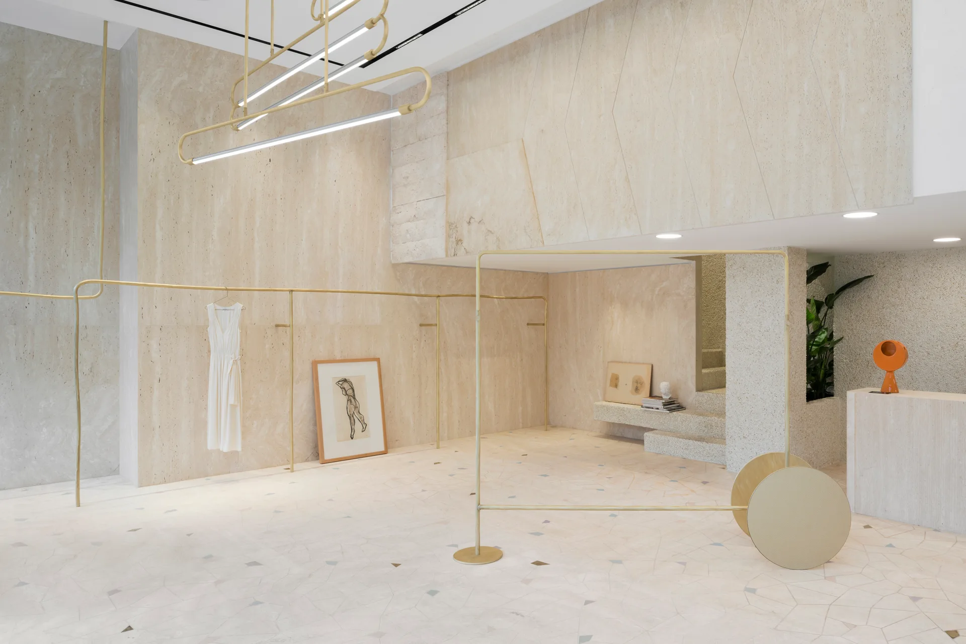 A minimalist retail space featuring textured travertine stone walls, slender brass clothing racks, and light-colored geometric stone tile flooring.