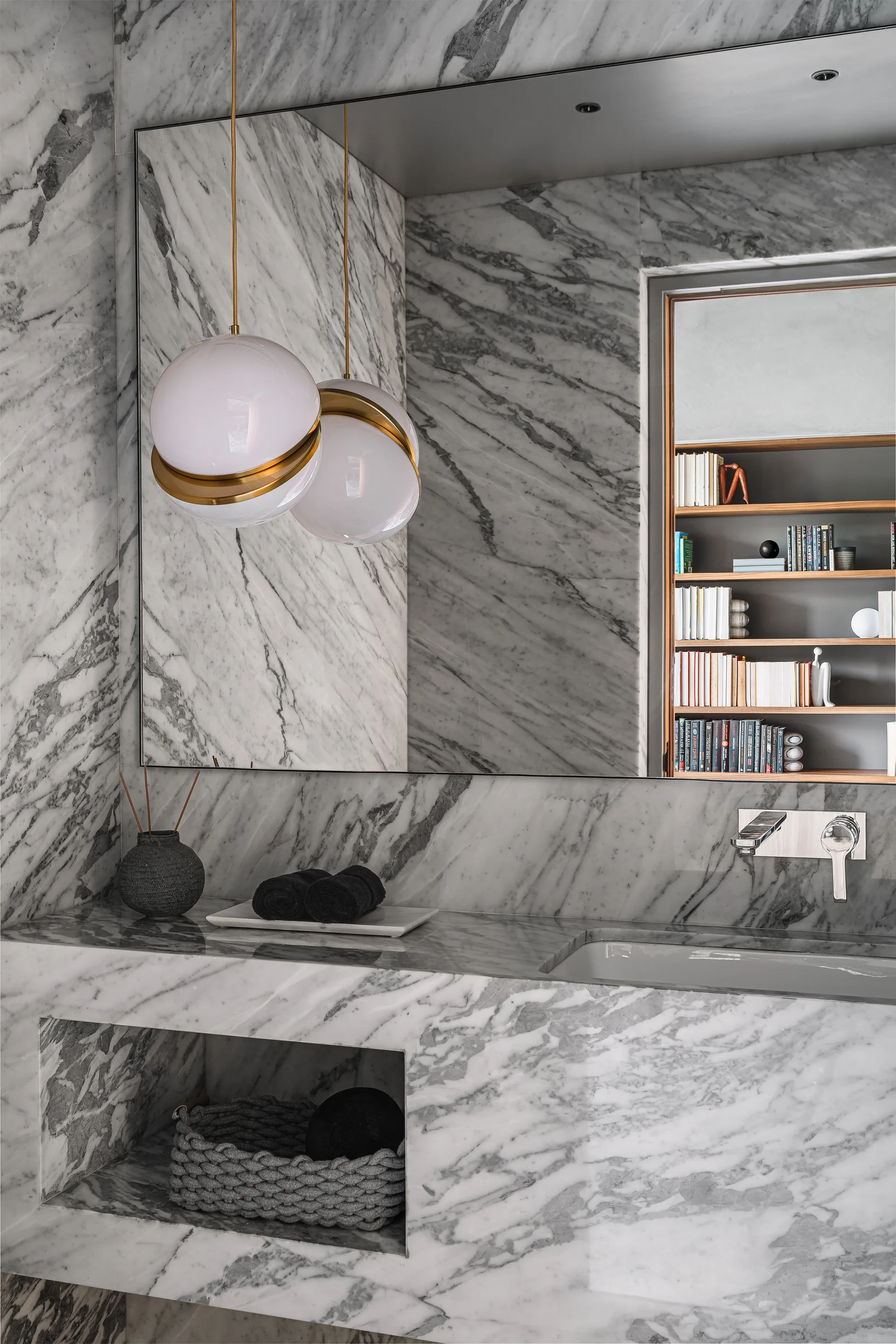 A marble-clad bathroom features an integrated stone vanity with a wall-mounted faucet, spherical pendant lights, and a large frameless mirror.