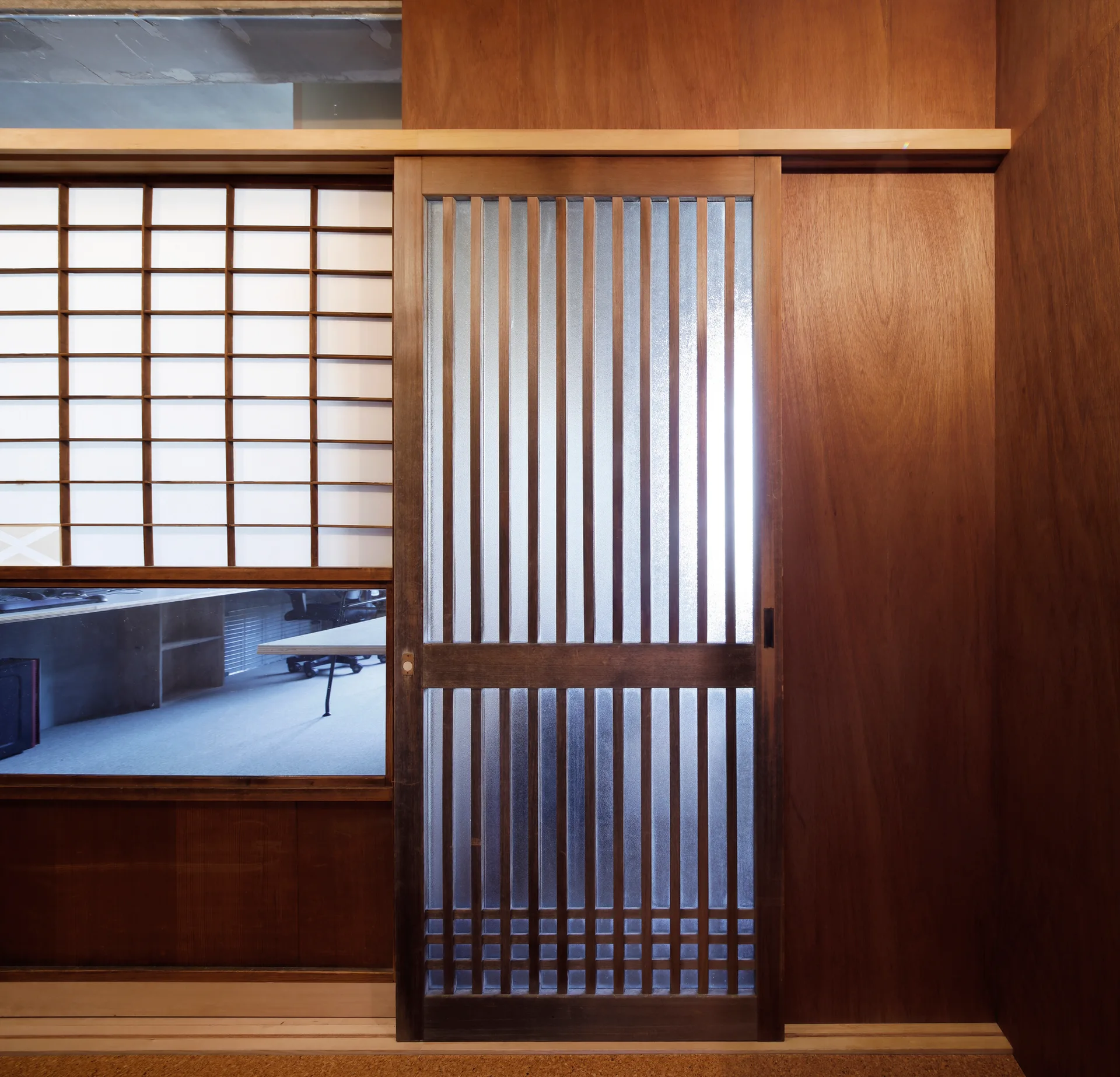 A wooden slatted sliding door with textured glass panels sits next to a grid-patterned screen and matching warm wood wall paneling.