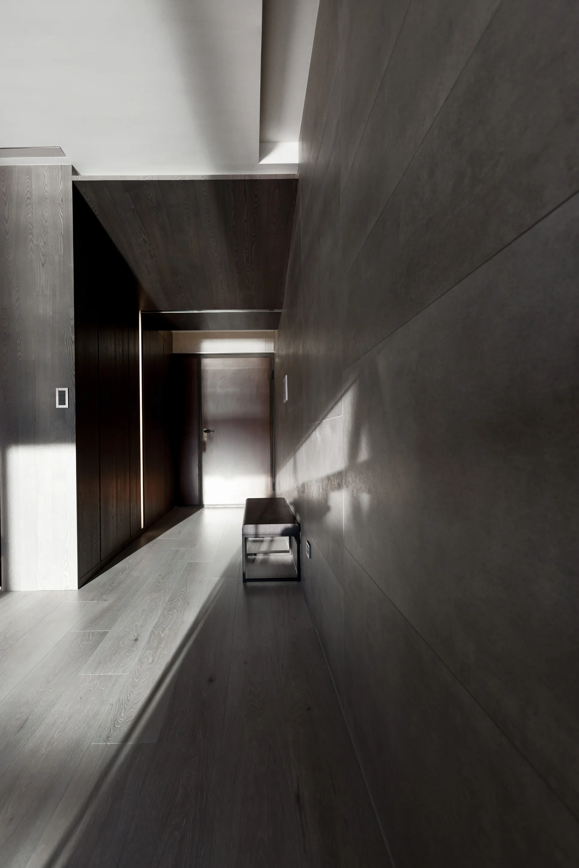 A long minimalist hallway features large dark gray wall panels, light wood plank flooring, and a single low bench in directional light.