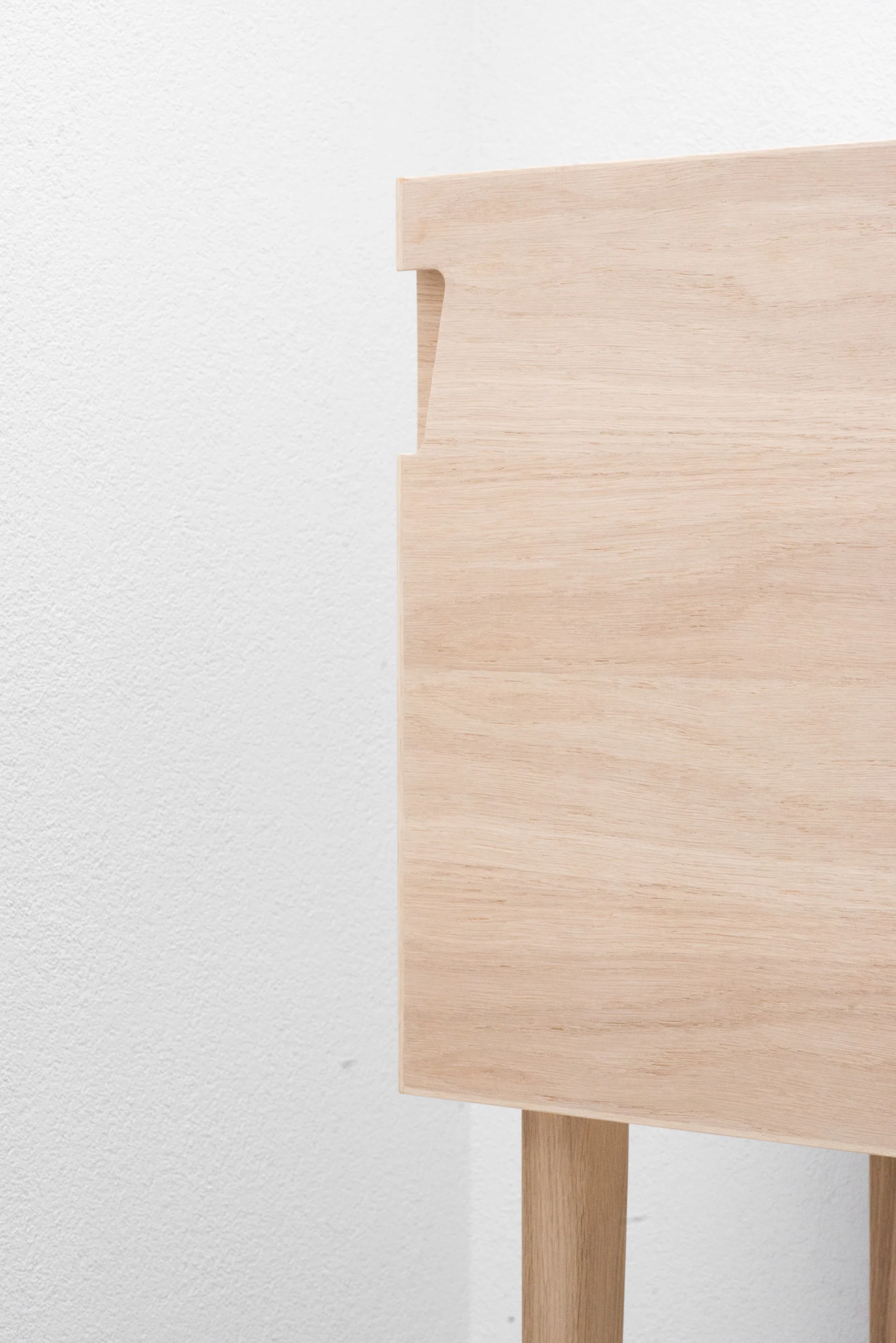 A close-up of a minimalist light wood cabinet featuring an integrated cutout handle and tapered leg against a textured white wall.
