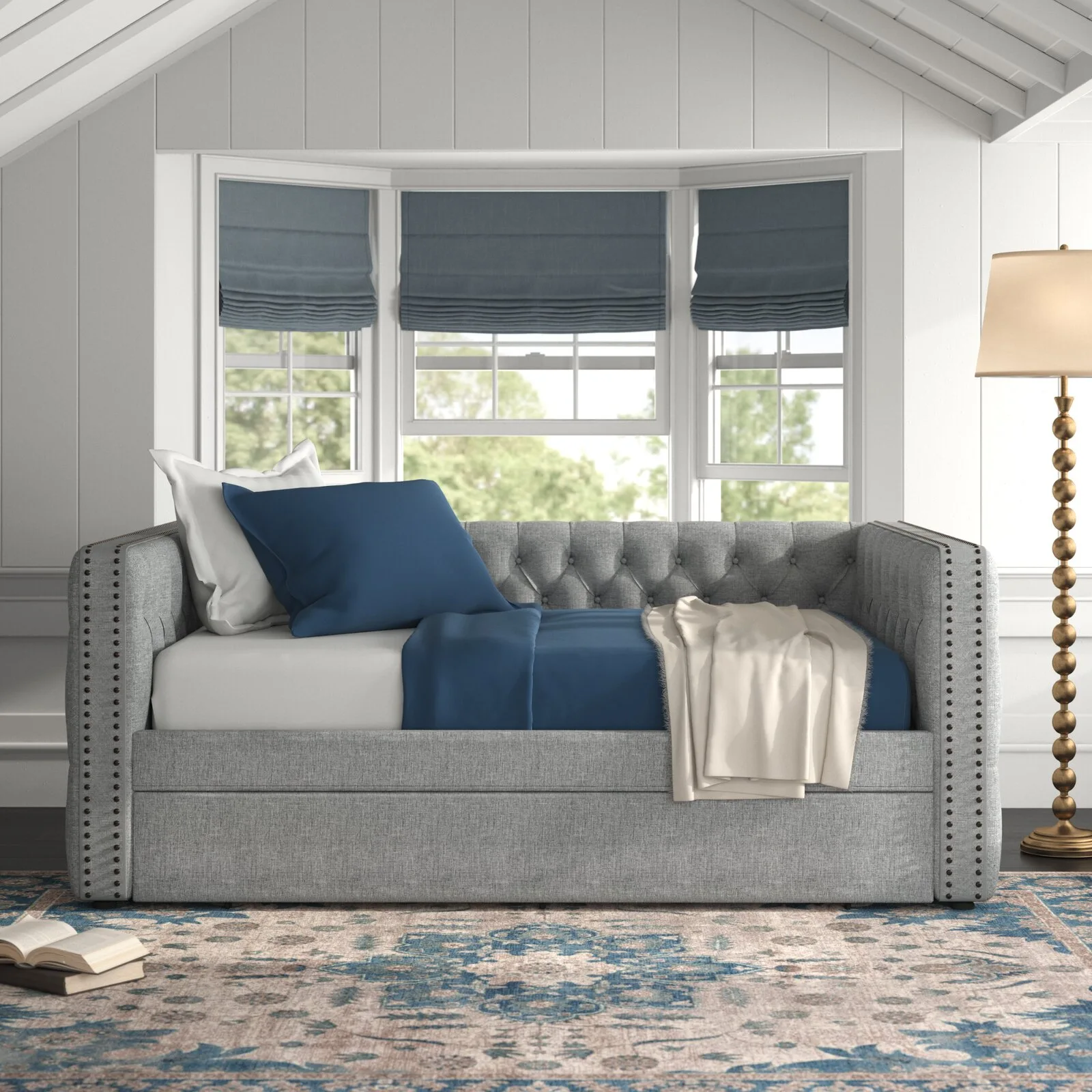 A grey tufted upholstered daybed with blue bedding sits under a bay window with matching blue roman shades in a room with white paneled walls.