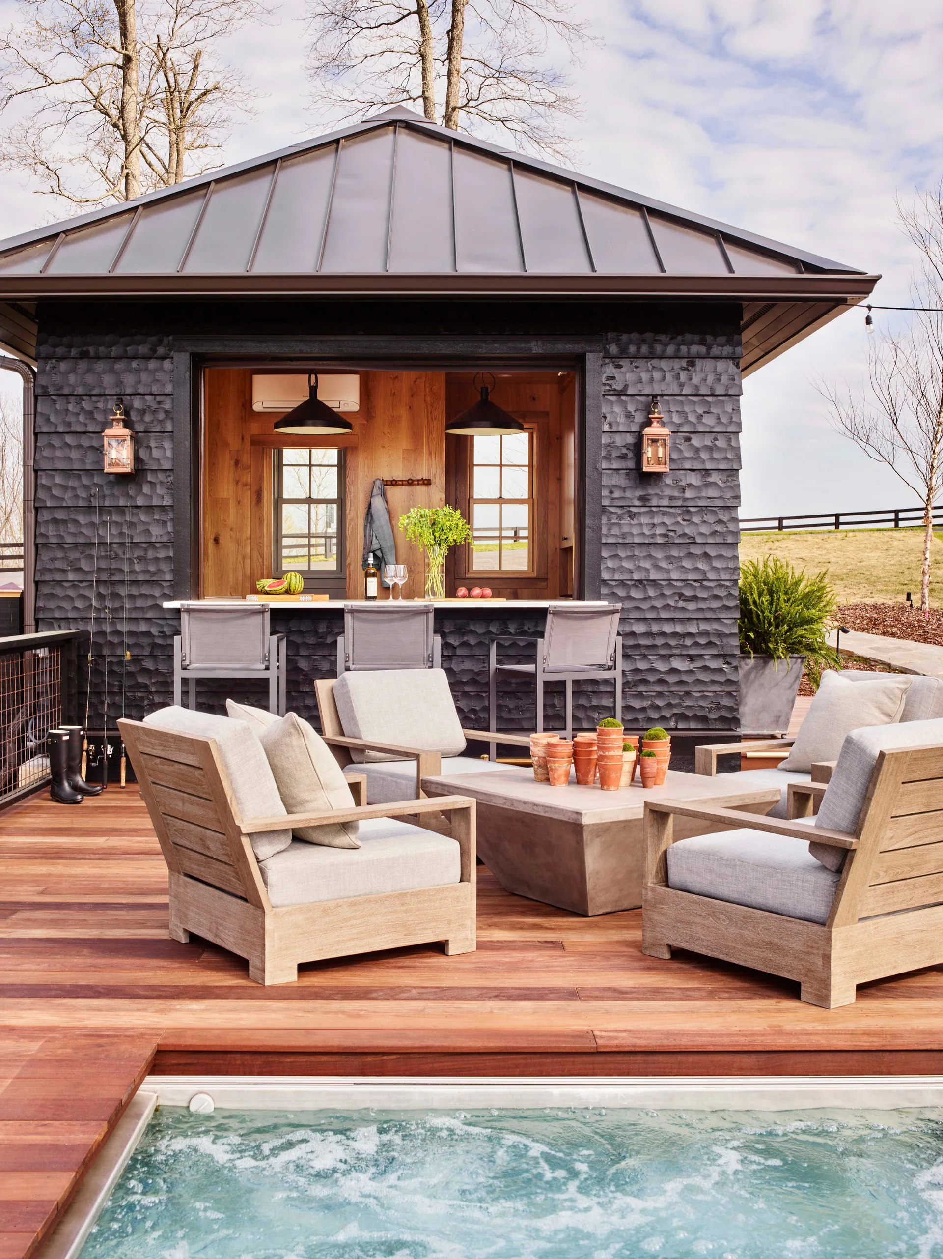 A wooden deck features a charcoal-shingle pool house with a pass-through bar, surrounding wood lounge chairs, and a spa in the foreground.