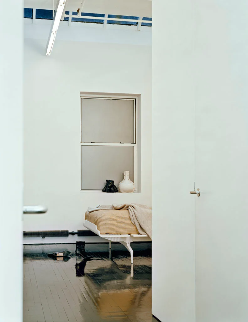A view through a white doorway reveals a low-profile bed, a window with a deep sill holding a vase, and dark reflective wood floors.