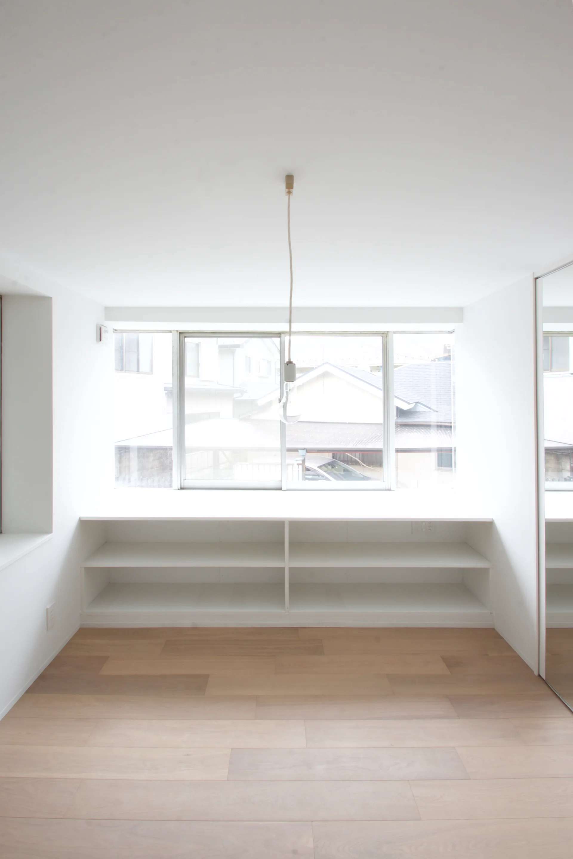 An empty white room features light wood plank floors, a large central window with low built-in shelving underneath, and a single hanging pendant light.