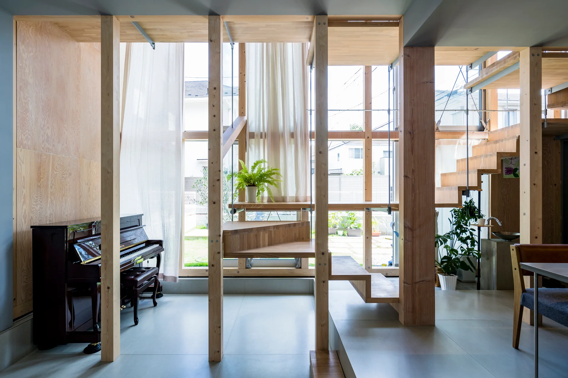 A modern wooden interior featuring exposed timber posts, a floating staircase, a black upright piano, and large windows looking out to a garden.