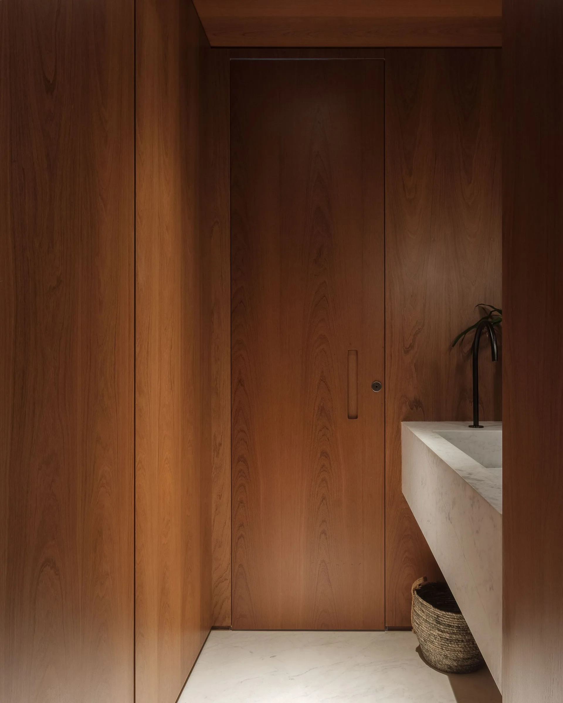 A minimalist bathroom features floor-to-ceiling vertical wood wall paneling, an integrated flush door, and a floating white marble vanity.