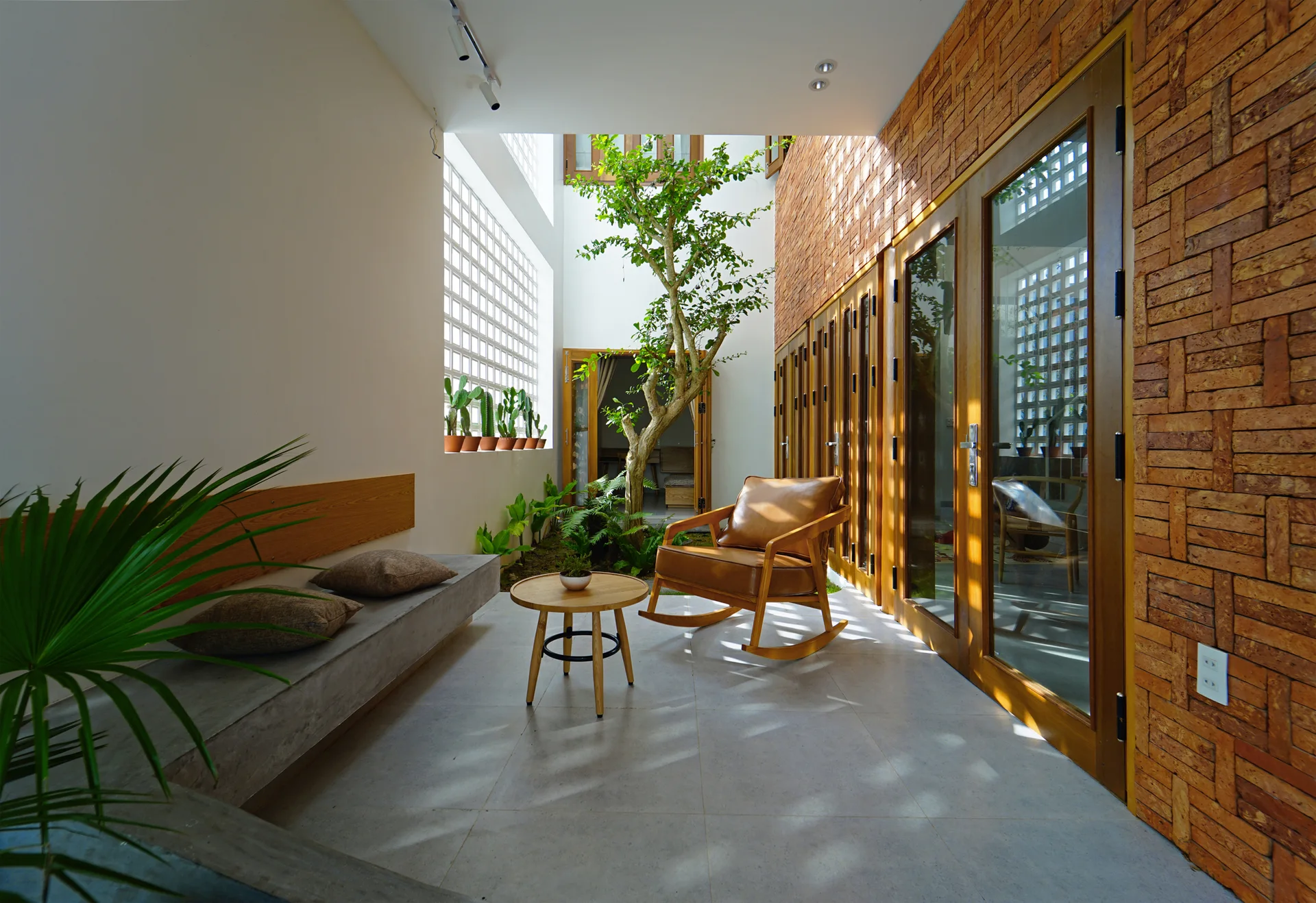 A modern sheltered courtyard featuring a central tree, a brick wall, wood-framed glass doors, a concrete bench, and a leather rocking chair.