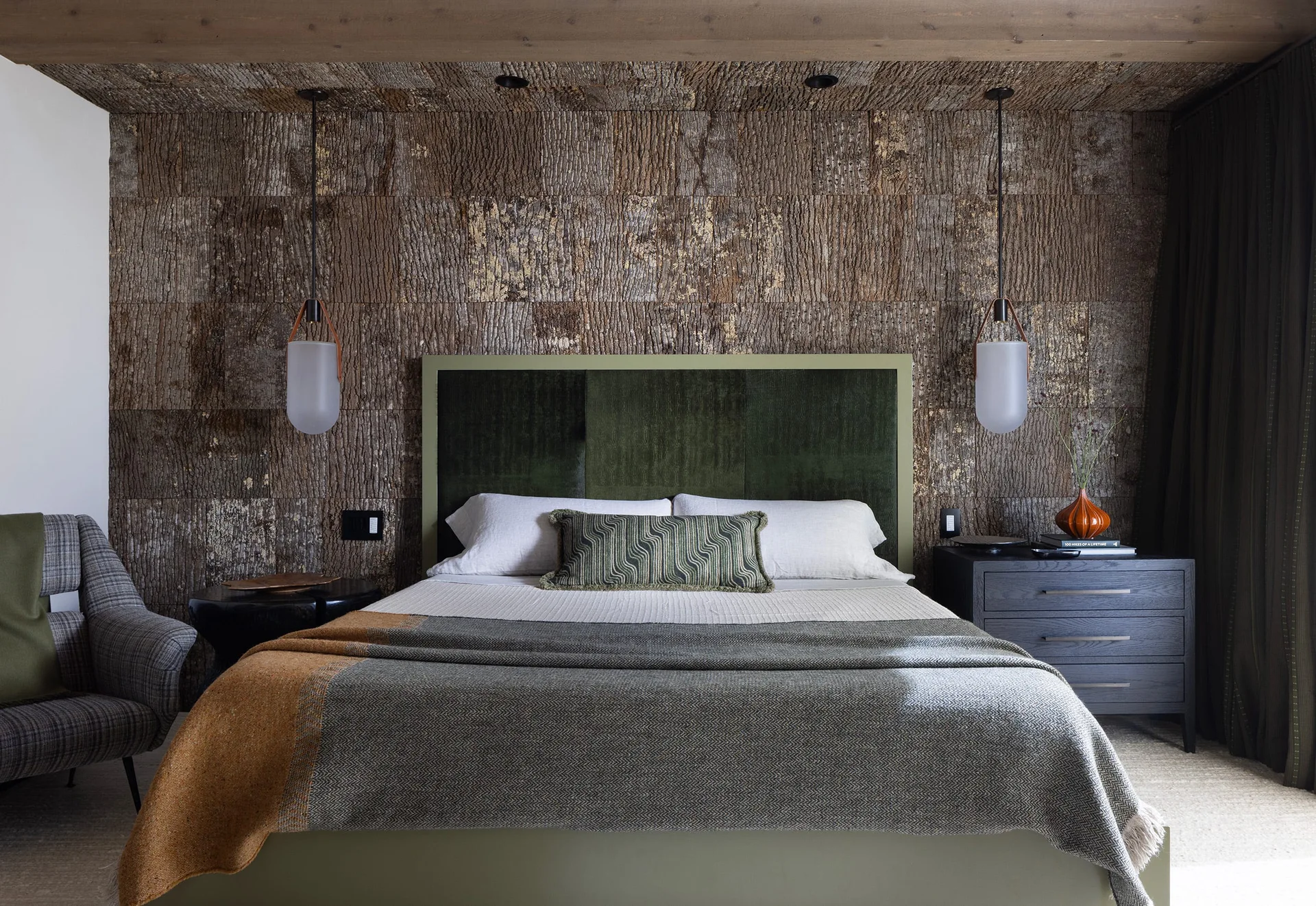 A modern bedroom features a sage green upholstered headboard against textured bark wall paneling flanked by frosted glass pendant lights.