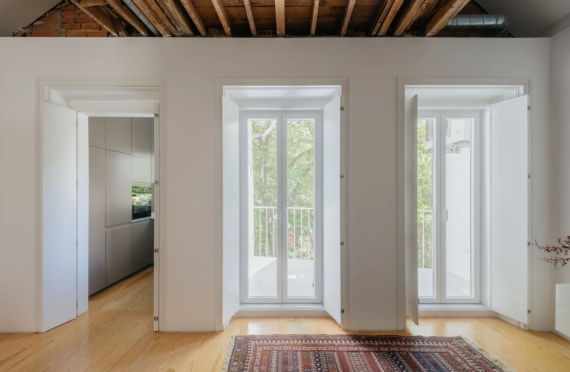 Bright interior with light wood flooring, an exposed timber ceiling, two tall white French windows, and an open doorway showing a gray kitchen.