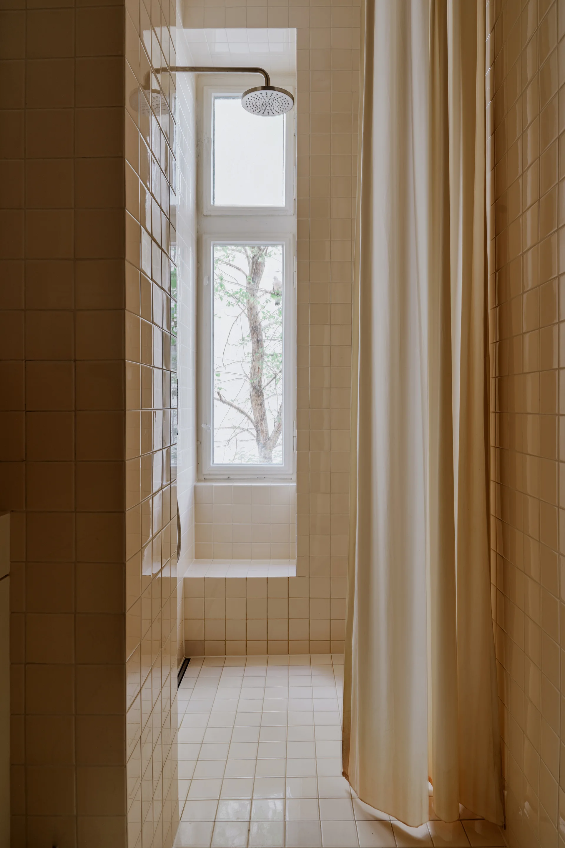 A shower stall features glossy beige square tiles on the walls and floor, a tall narrow window, a metal showerhead, and a cream curtain.