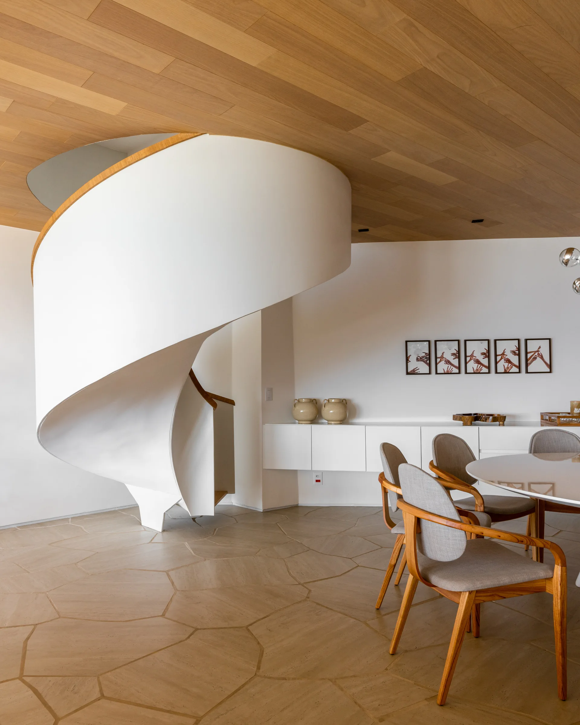 A modern dining area featuring a prominent white spiral staircase, wood plank ceiling, stone tile flooring, and a dining table with wooden chairs.