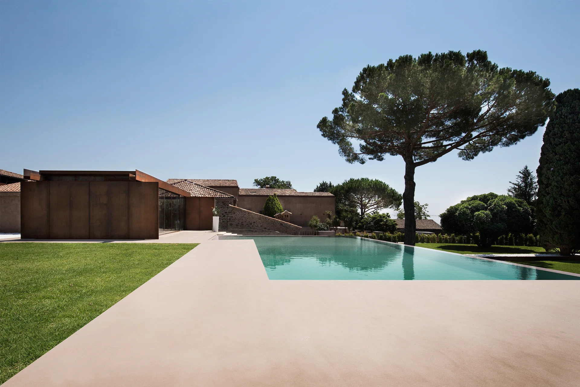 A rectangular swimming pool is flanked by a tan concrete deck, manicured green lawn, and a modern building with rust-colored metal panels.