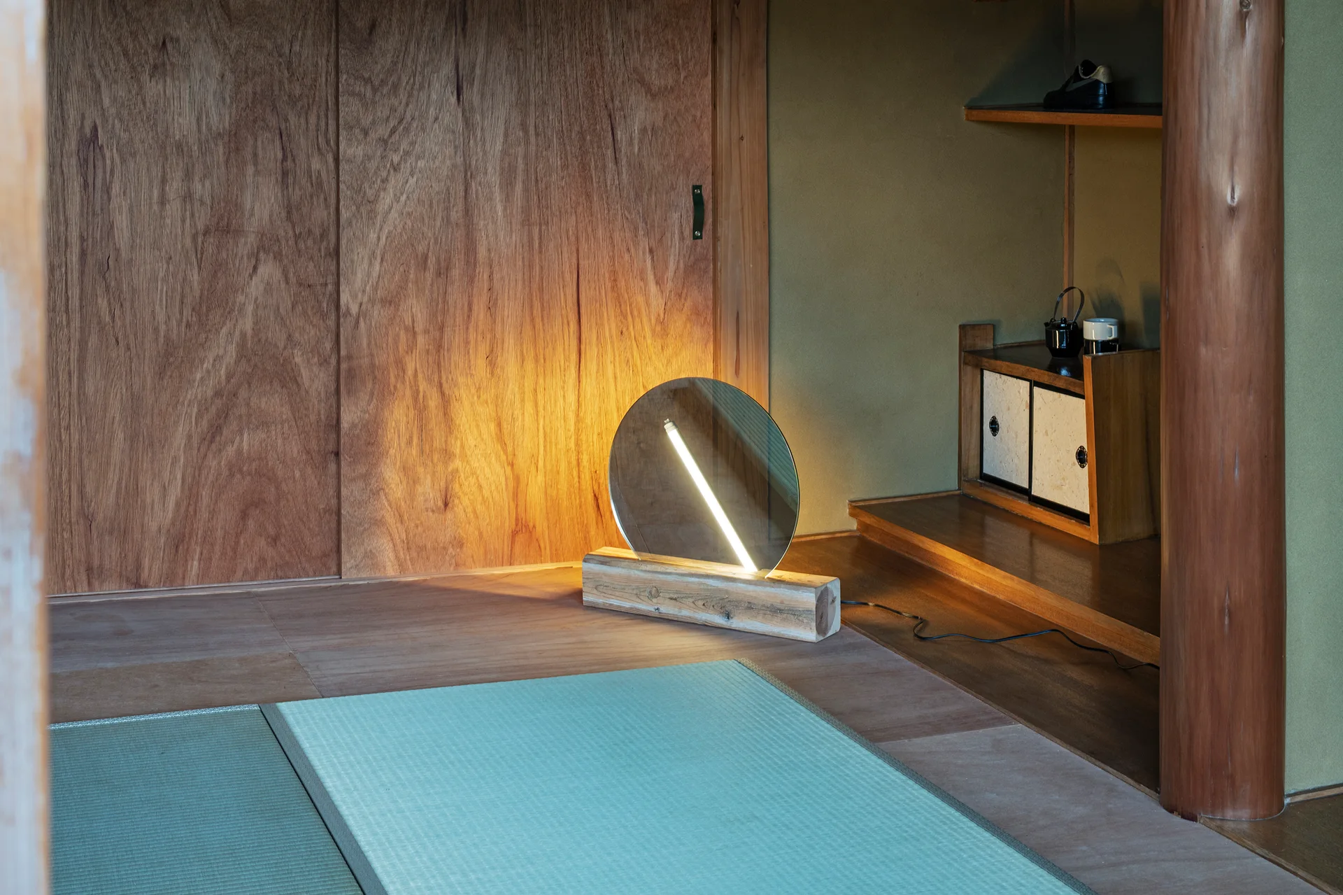 A minimalist circular mirror lamp glows on a wooden block next to light green tatami floor mats and a large wood grain sliding door.