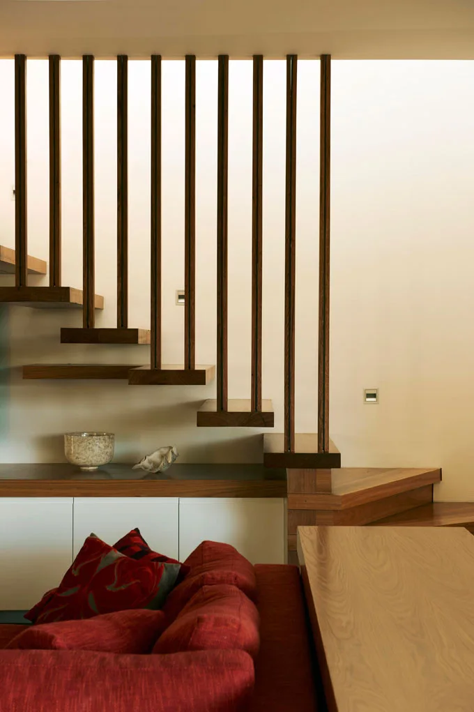 A modern floating wood staircase features vertical wooden slat railings, set behind a red sofa and a white storage cabinet.