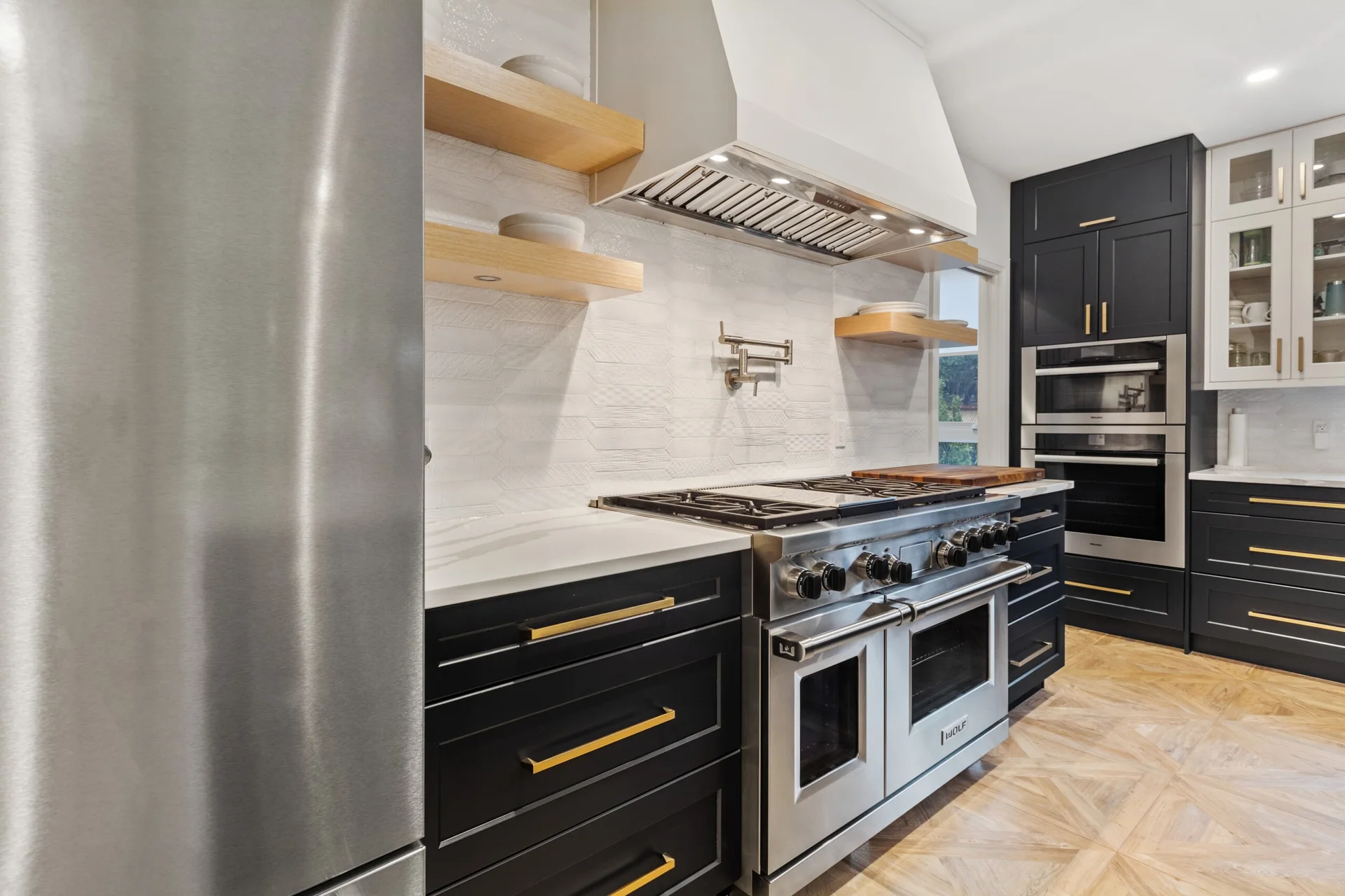 A modern kitchen featuring dark charcoal lower cabinets with gold handles, light wood floating shelves, a white tiled backsplash, and a stainless steel range.