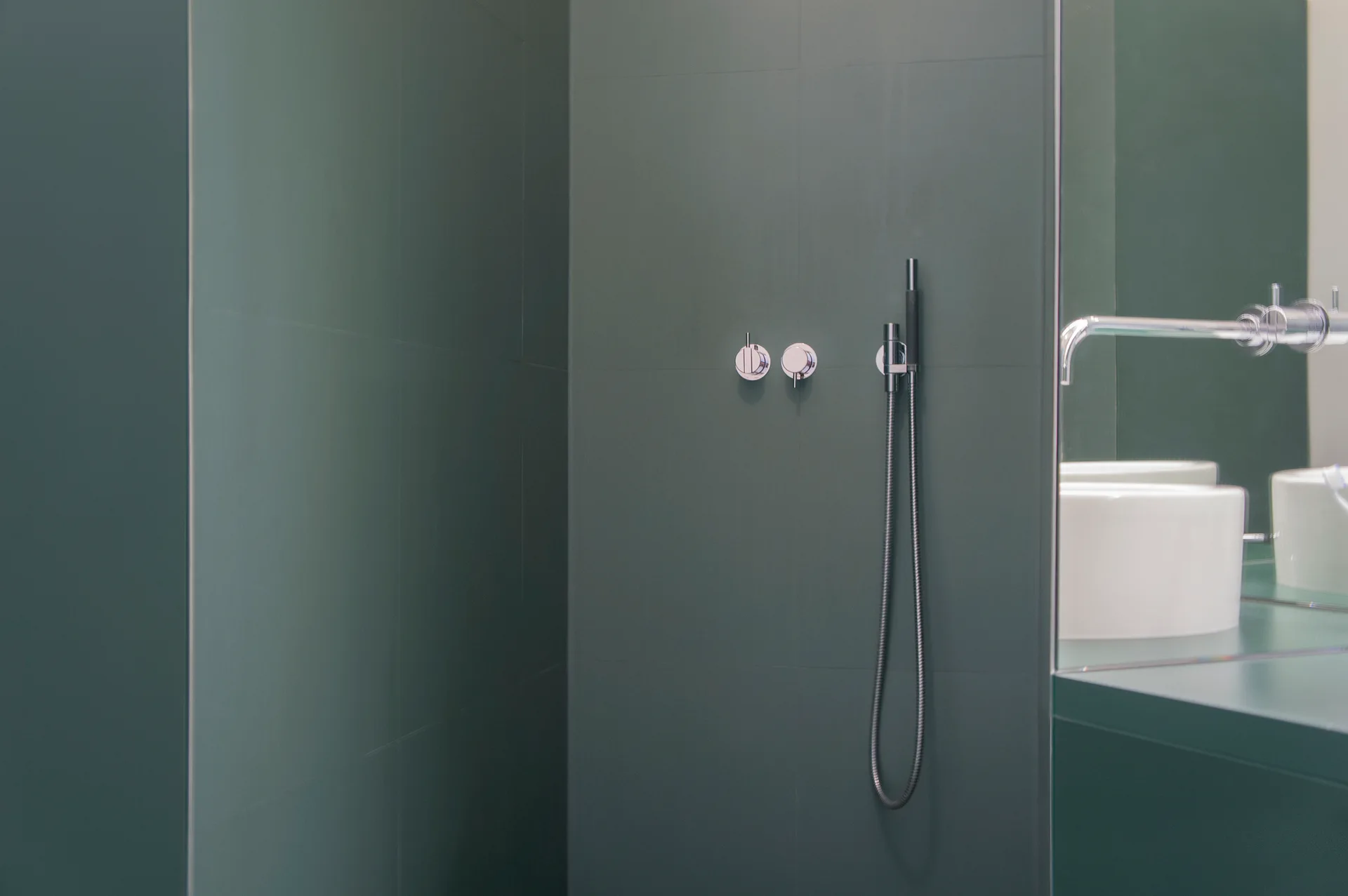 A dark green tiled shower wall features minimalist chrome controls and a handheld wand, next to a mirror reflecting a white vessel sink.