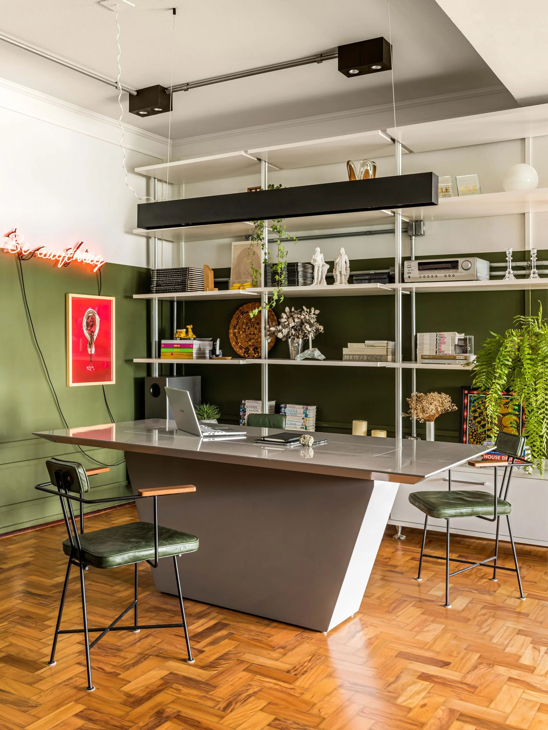 A home office featuring a large grey desk, green leather chairs, and a floor-to-ceiling metal shelving unit against a deep green wall.