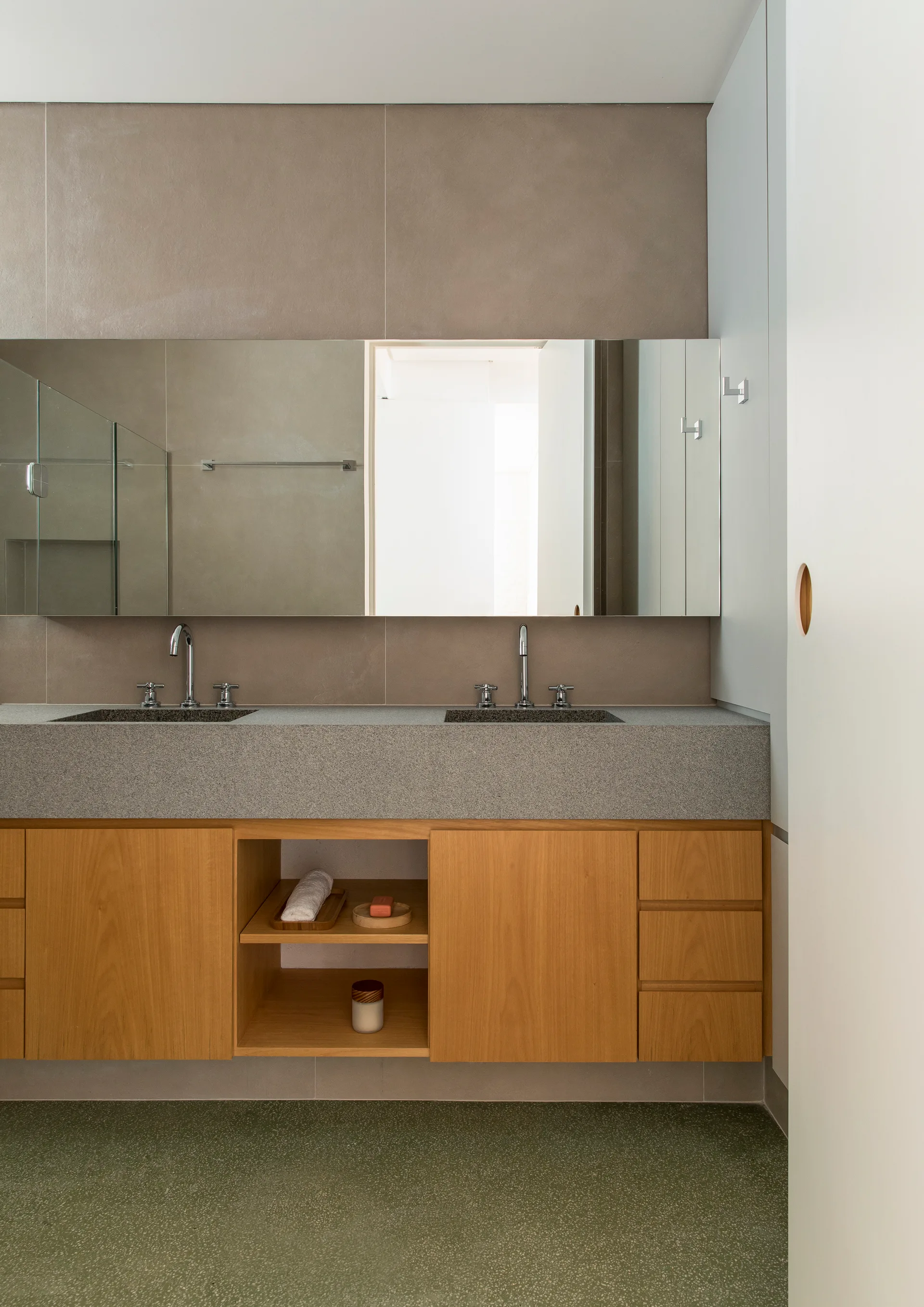A minimalist bathroom with a wooden double vanity, grey stone countertop, integrated sinks, wide wall mirror, and green speckled floor.