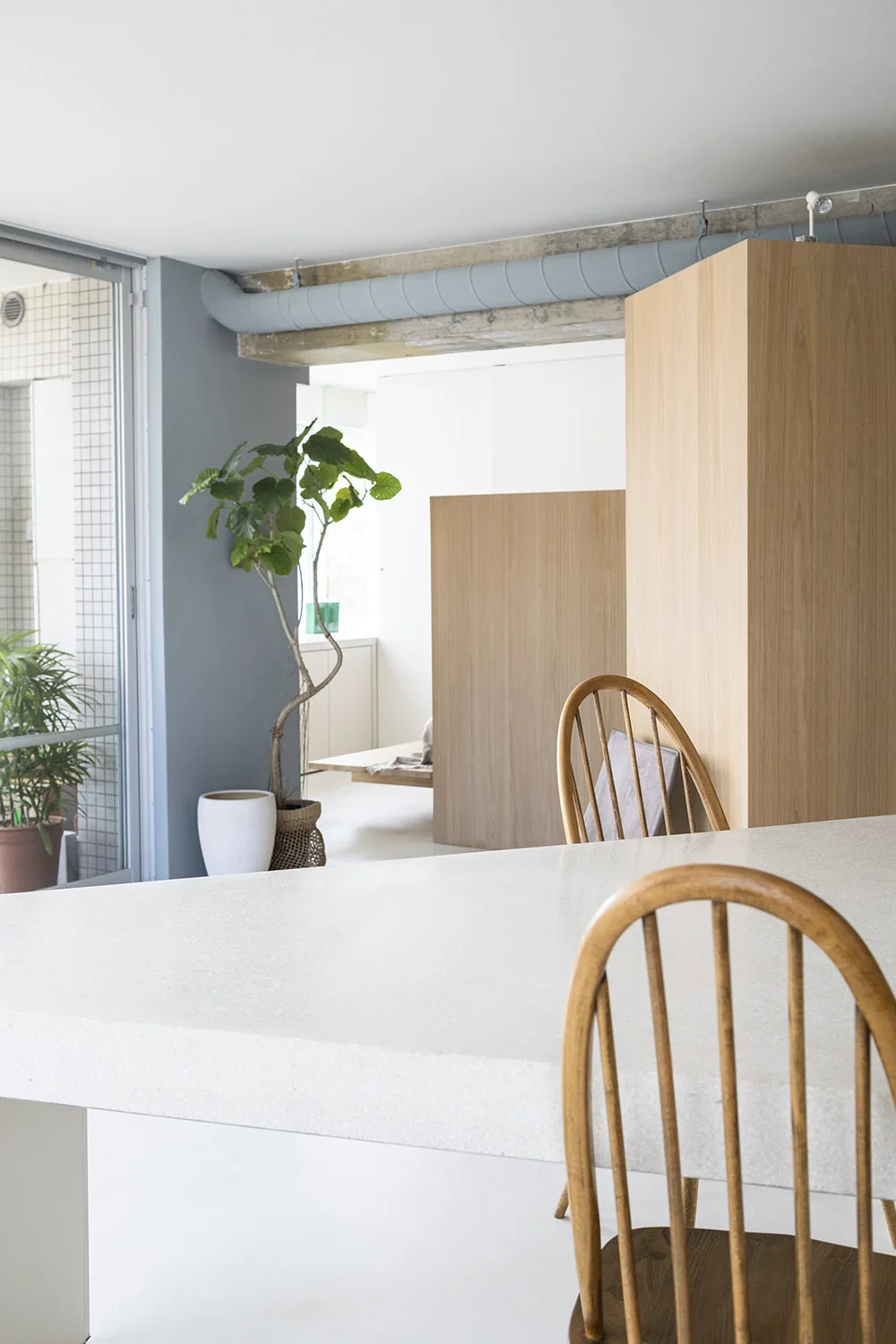 A minimalist dining area featuring a speckled white stone table, wooden spindle-back chairs, a tall oak cabinet, and an exposed ceiling duct.