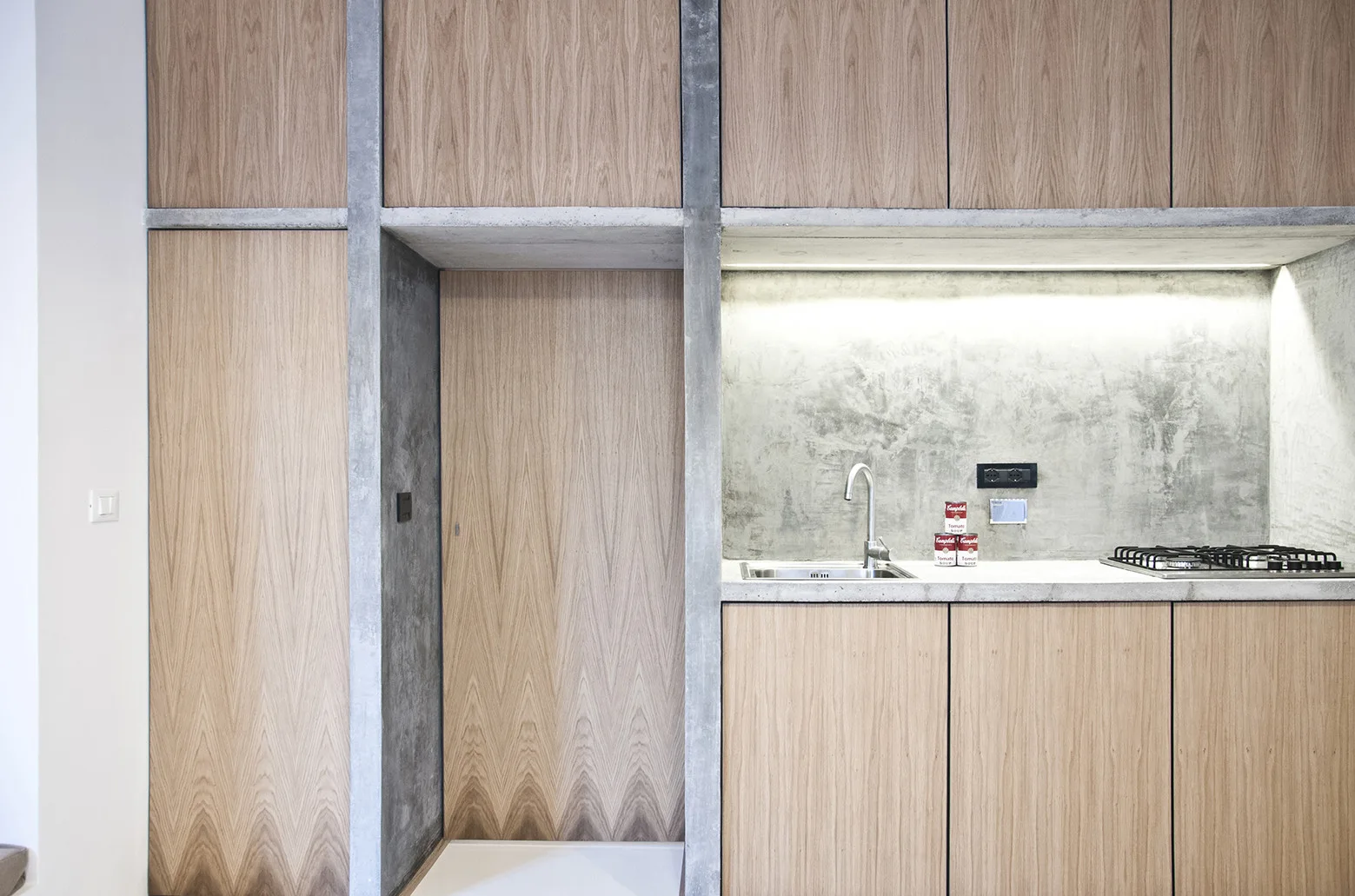 A minimalist kitchen featuring light wood cabinets framed by exposed concrete columns, a concrete countertop with a sink, and integrated under-cabinet lighting.